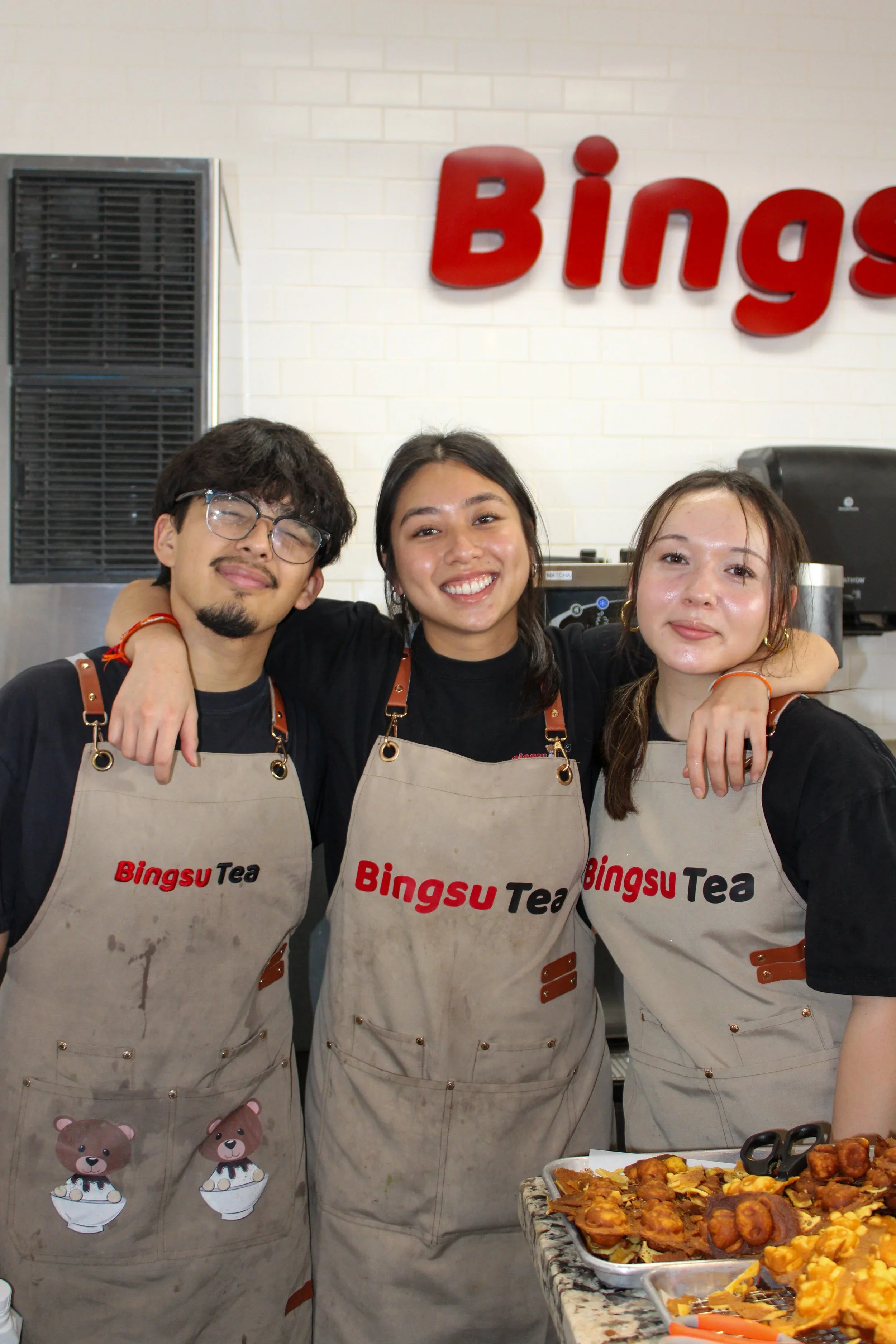 Three smiling people in aprons with 'Bingsu Tea'. The person on the left is a man with glasses and a beard, the middle person is a woman with long dark hair, and the person on the right is a woman with light brown hair. There are food trays with snacks on the right, and a white brick wall with red lettering in the background.