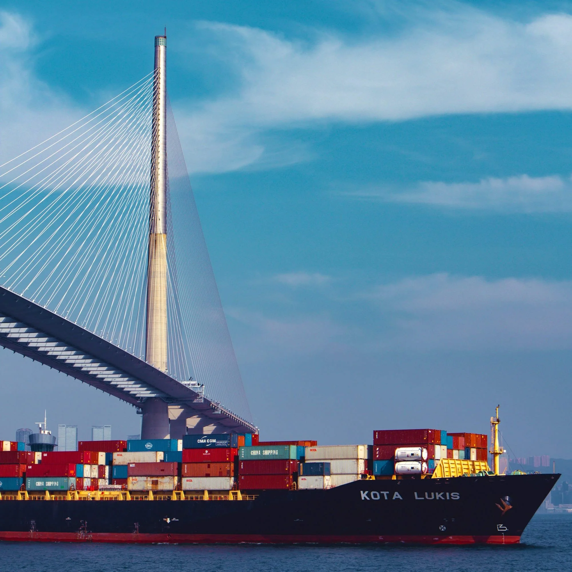 A large cargo ship loaded with colorful shipping containers sailing in the water with a modern cable-stayed bridge and city skyline in the background.