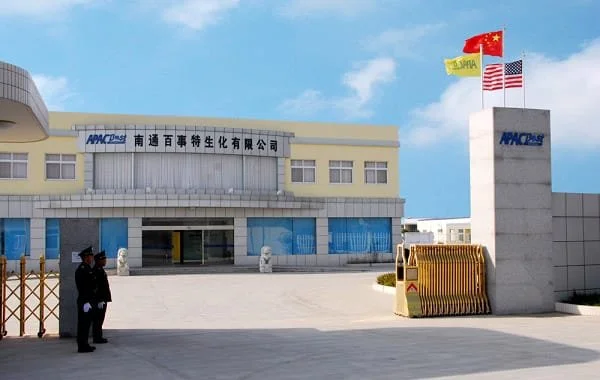 Entrance of a company building with flags of China, the USA, and a yellow flag, with a guard standing near a barrier.