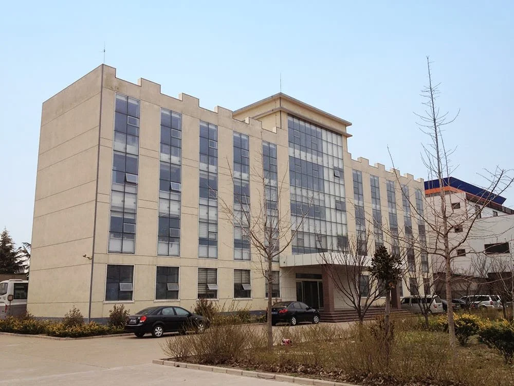 A multi-story beige office building with glass windows and a parking lot in front, surrounded by leafless trees.