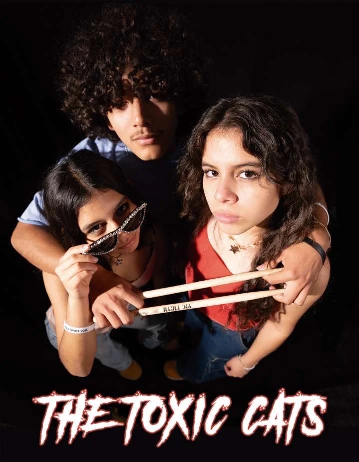 Three young people looking up at the camera, two women and one man, with serious expressions. One woman is holding a pair of drumsticks and sunglasses. The background is black, and the text 'THE TOXIC CATS' appears in stylized red and white letters at the bottom.