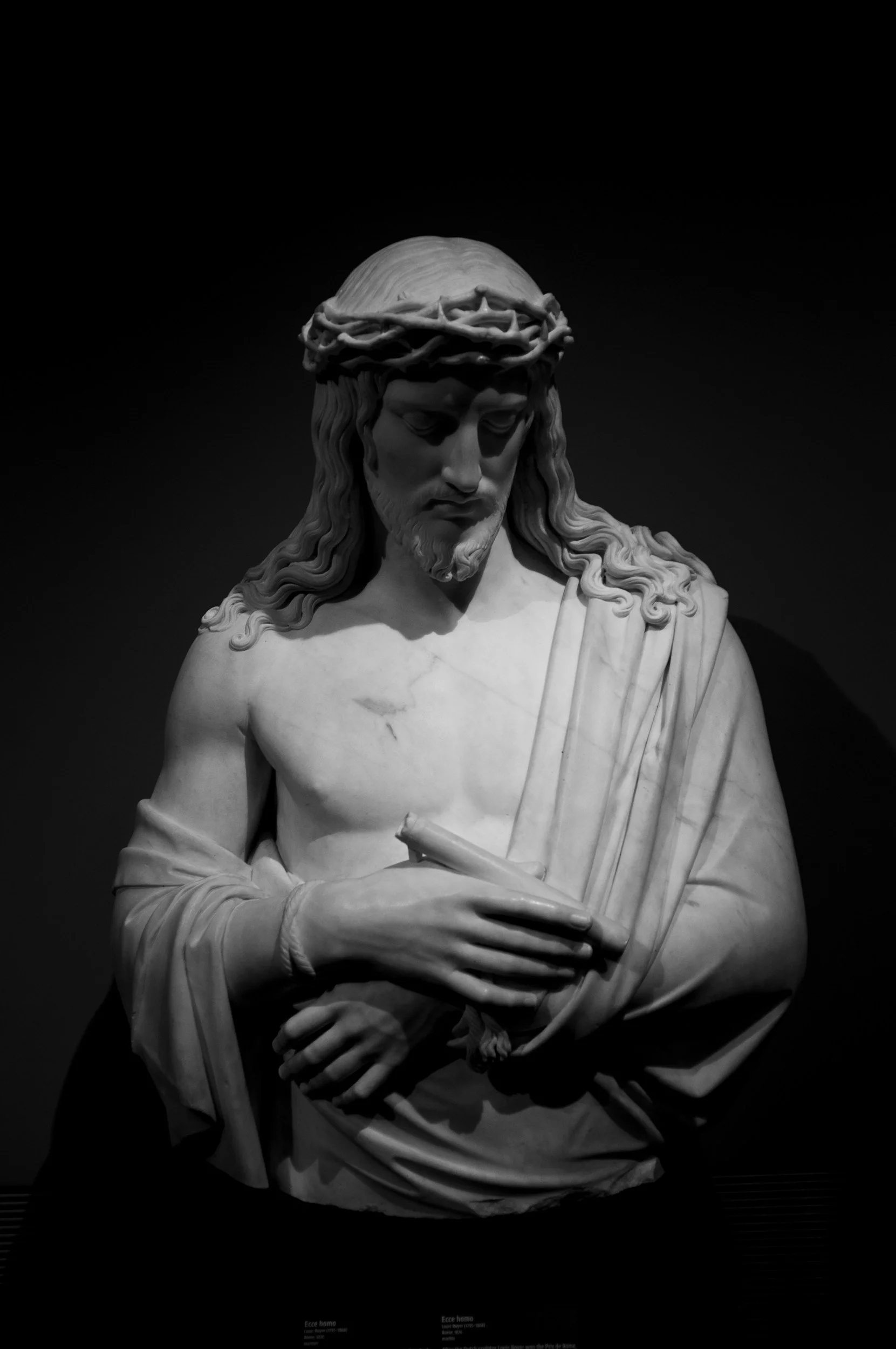 Black and white photograph of a marble bust of Jesus Christ, wearing a crown of thorns, with downward gaze, draped in robes, holding a scroll in his left hand against a dark background. (Not my original work)