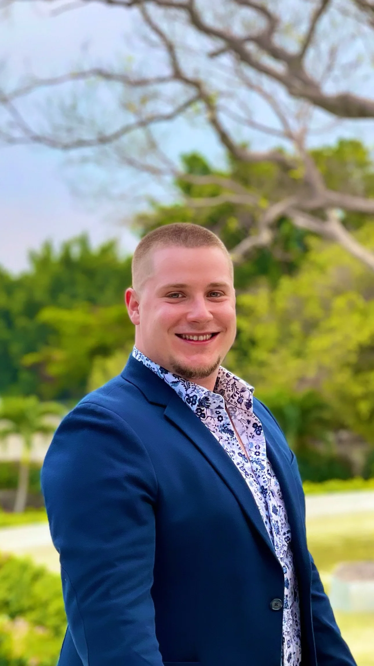 Portrait of a young man with short light brown hair and a goatee, smiling, wearing a blue blazer and a patterned shirt, outdoors with green trees and cloudy sky in the background.