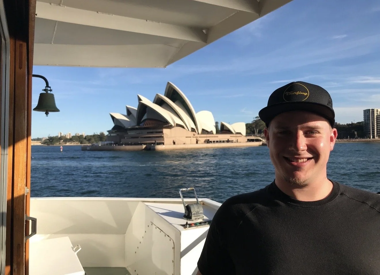 A smiling man wearing a black cap and shirt standing on a boat, with the Sydney Opera House and Sydney Harbour in the background during daytime.