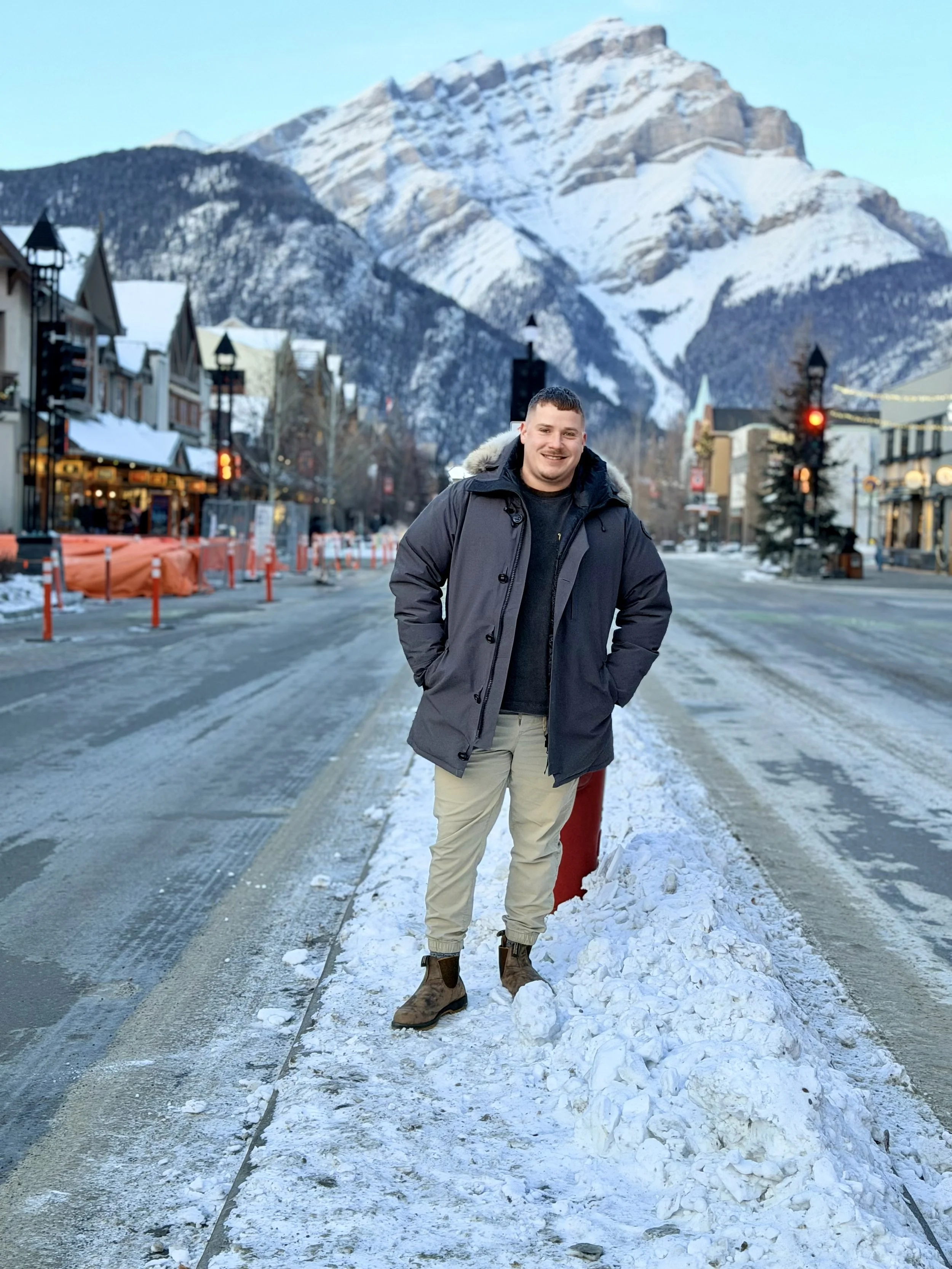 A smiling man in a black winter jacket and beige pants standing on a snowy street with a mountain in the background, under a clear sky.