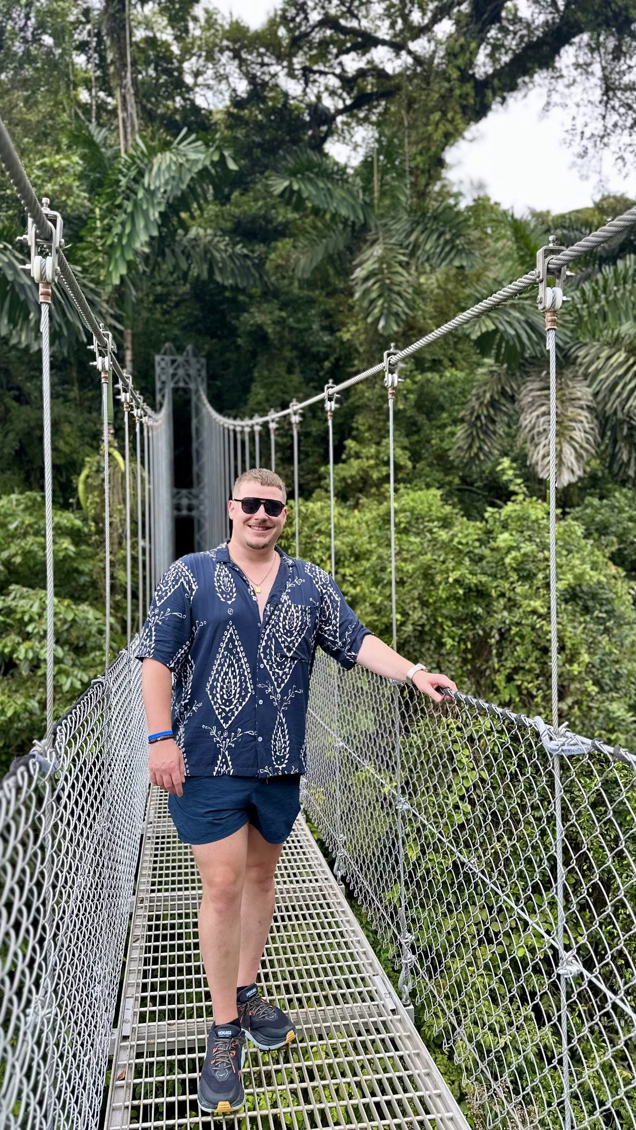 A man wearing sunglasses, a blue patterned shirt, and shorts standing on a suspension bridge amid lush green trees.