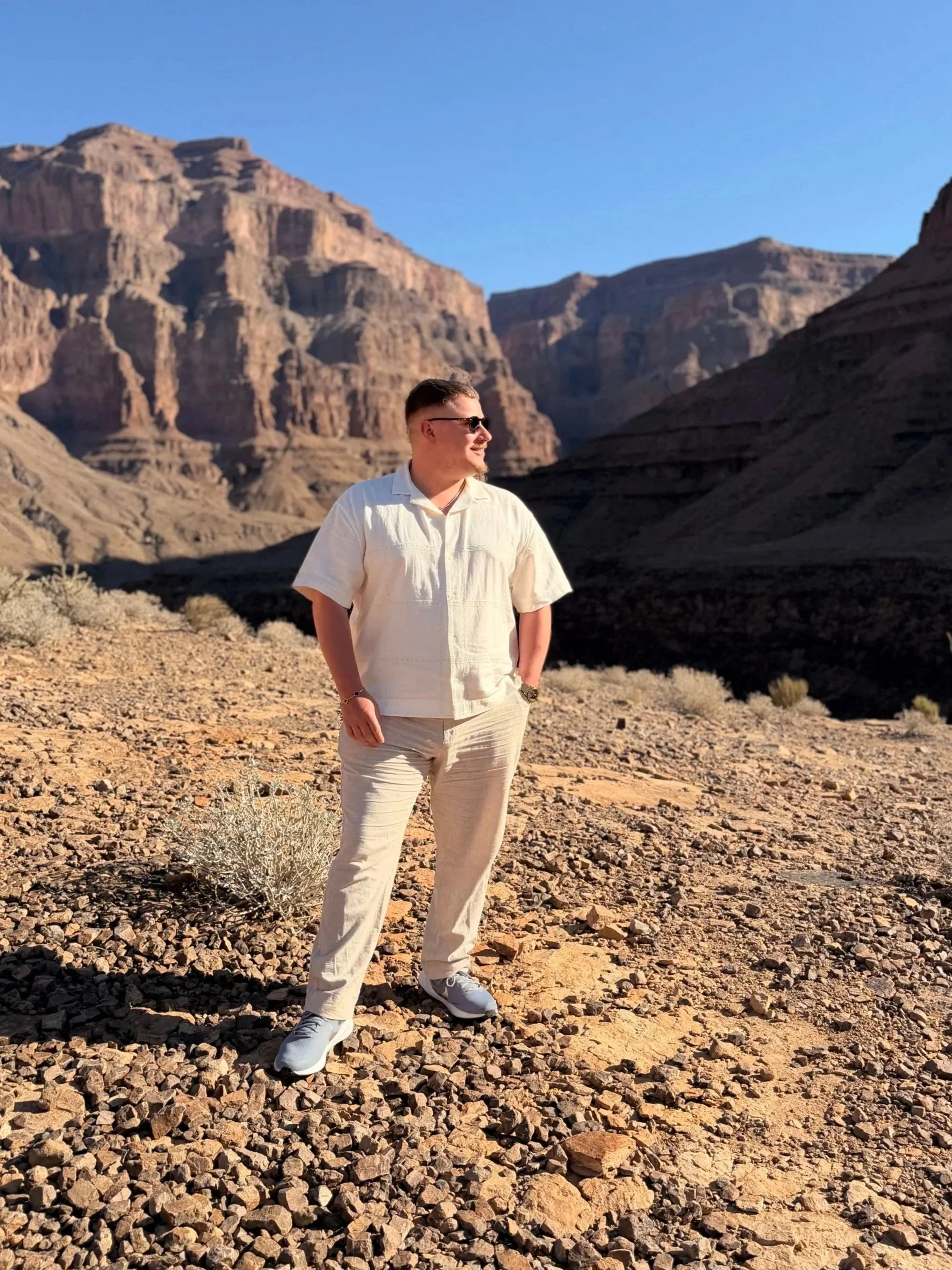 A man wearing sunglasses, a white shirt, and beige pants stands in a rocky desert landscape with large canyon walls in the background under a clear blue sky.