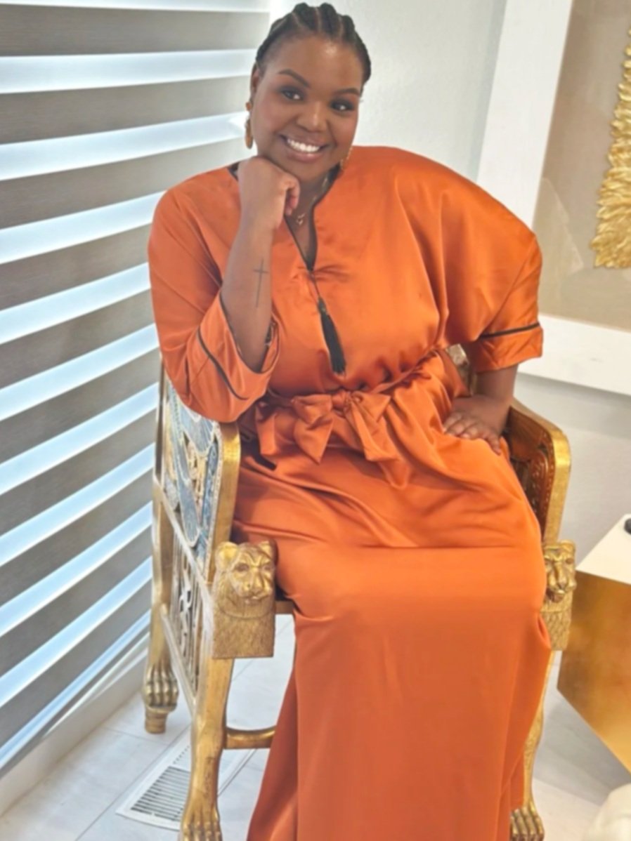 A woman wearing an orange satin dress with a waist tie, sitting on a ornate gold and carved wooden chair, smiling with her hand resting on her chin.