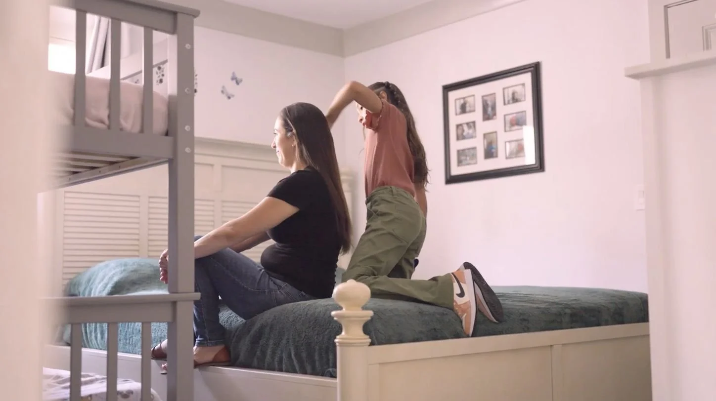 Two women on a bed in a bedroom, one sitting and the other kneeling, interacting near the head of the bed. The room has white walls, framed photos on the wall, and a bunk bed partially visible.