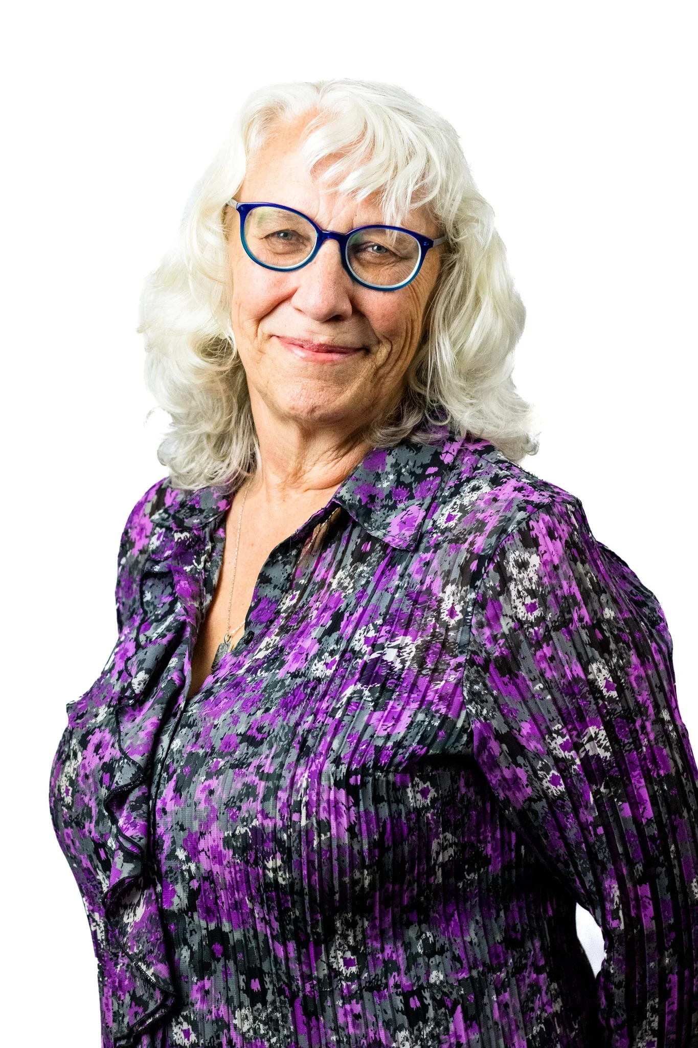 Senior woman with curly white hair and blue glasses, wearing a purple, black, and gray patterned blouse, smiling against a white background.