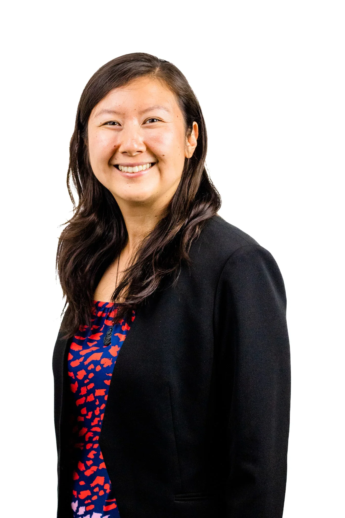 A smiling woman with long dark hair wearing a black blazer over a colorful patterned top, standing against a white background.