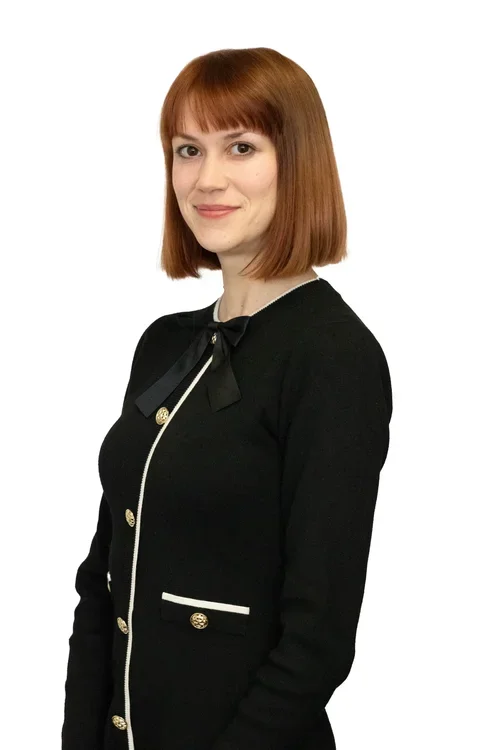 Portrait of a woman with long dark hair, smiling, wearing a black blazer, a polka dot blouse, pearl necklace, and makeup, against a white background.