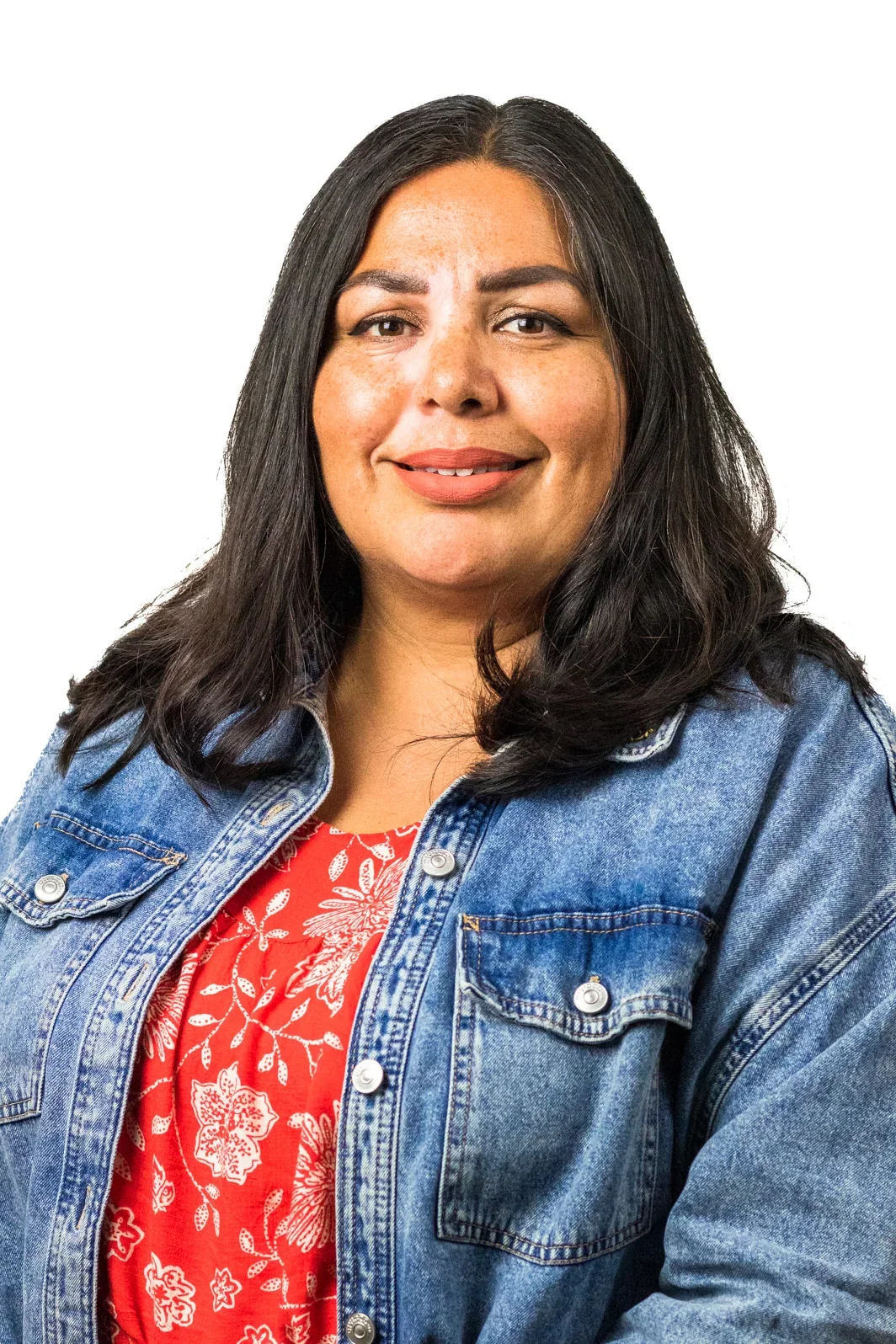 A woman with medium skin, dark wavy hair, and brown eyes, smiling, wearing a denim jacket over a red floral top, against a white background.