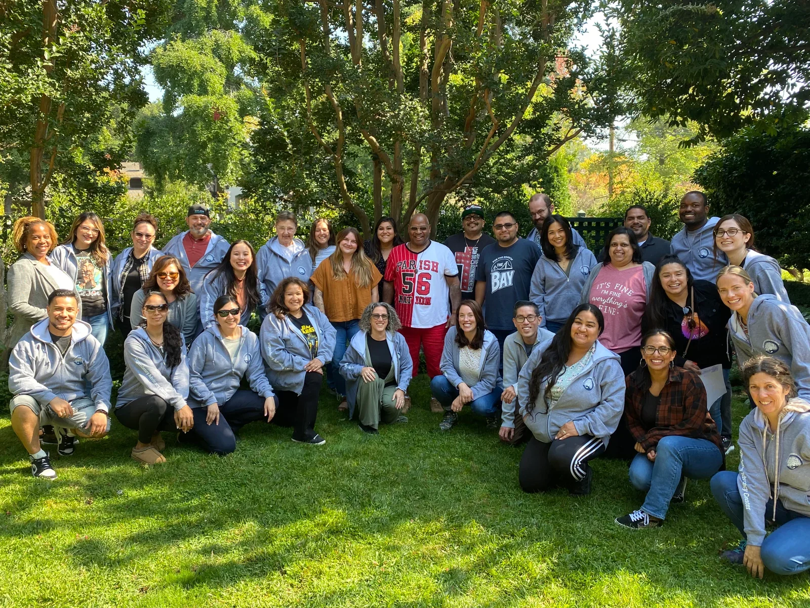 Group of diverse people posing for a photo outdoors in a green park, many wearing gray jackets.