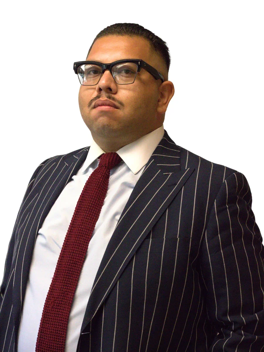 A man dressed in a dark pinstripe suit, white shirt, and red tie, wearing black glasses, standing against a white background, looking confident.