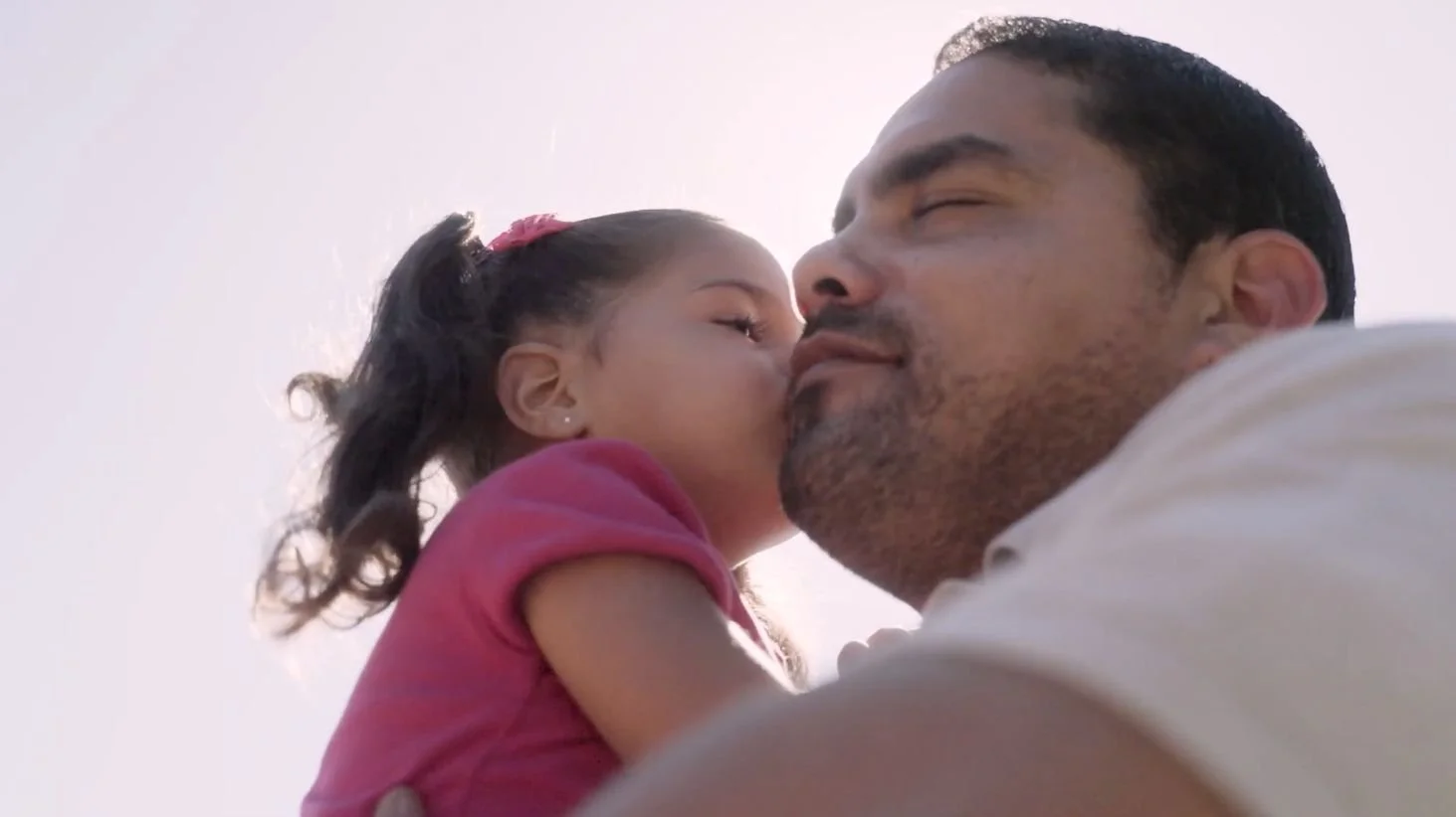 A young girl in a pink shirt kisses a man on the cheek outdoors on a sunny day.