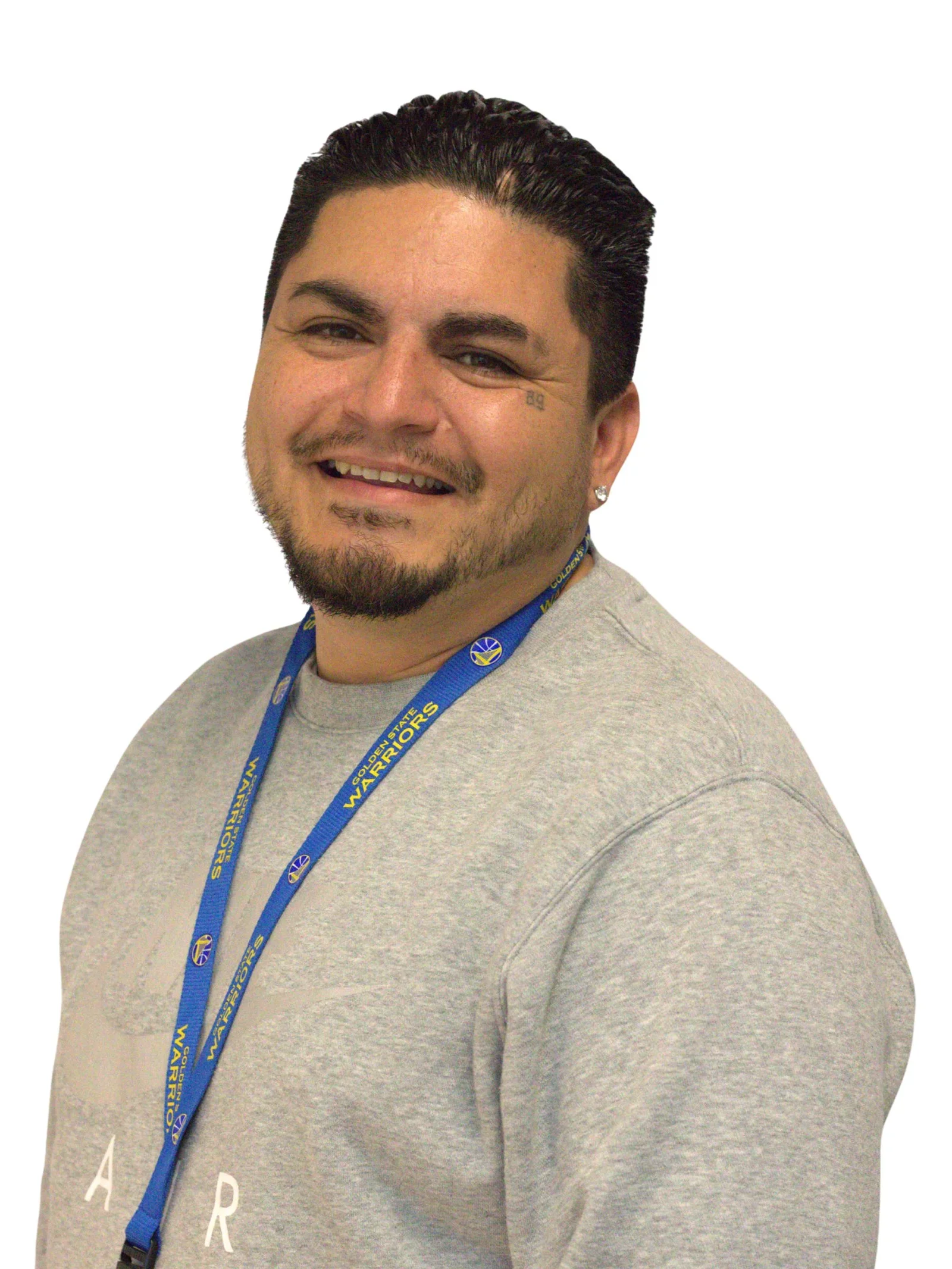 Close-up of a smiling man with dark slicked-back hair, facial hair, and a earring, wearing a gray shirt and a blue Golden State Warriors lanyard.