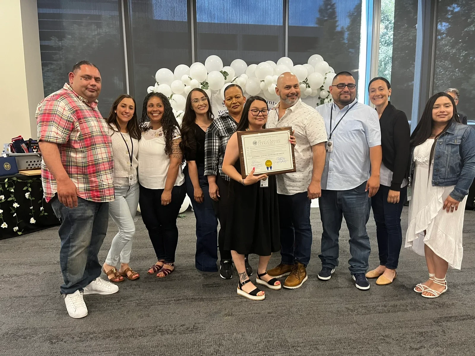 A group of people posing with a framed certificate at Santa Clara County's 14th Annual Reunification Day Celebration, with a balloon arch in the background.