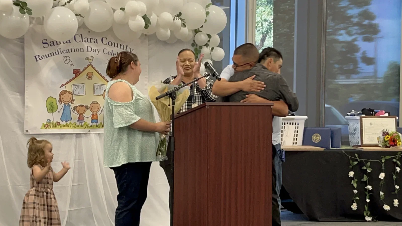People embracing on stage at Santa Clara County's 14th Annual Reunification Day Celebration, with a banner and balloon arch in the background.