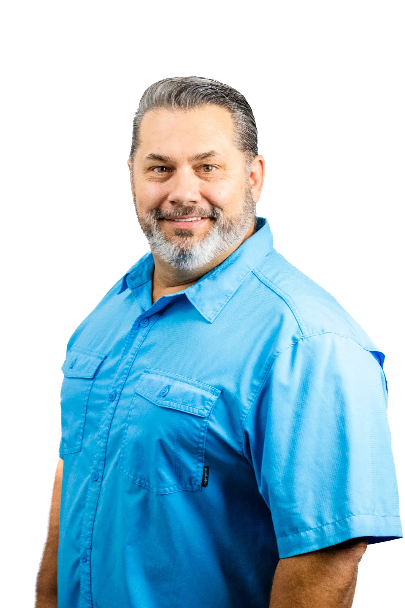 A middle-aged man with gray hair and a beard, smiling and wearing a light blue short-sleeved button-up shirt, against a white background.