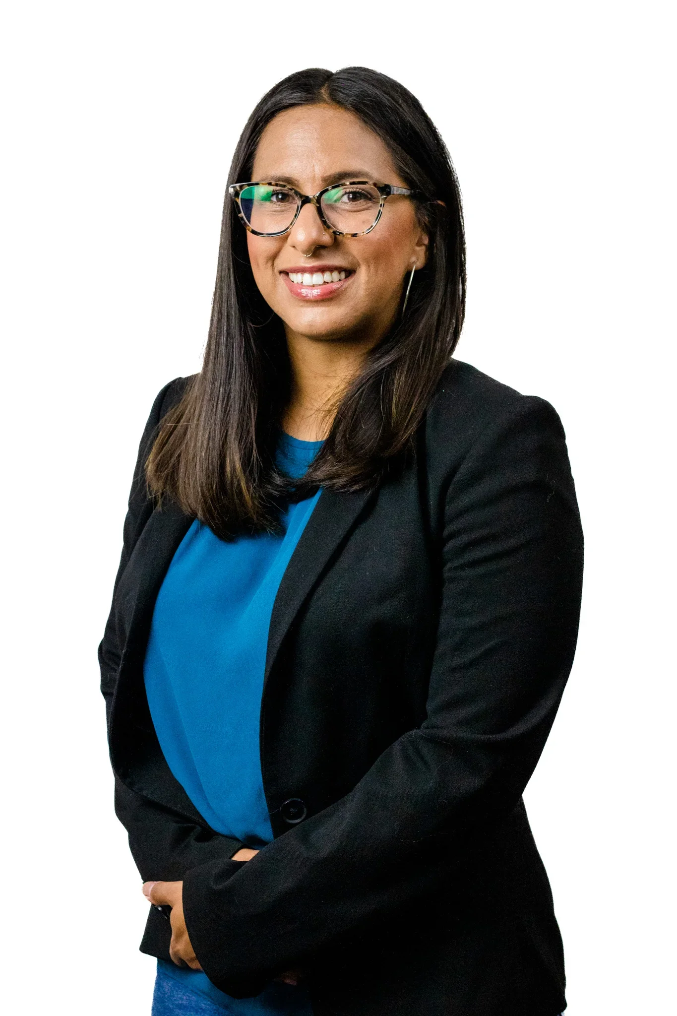 A woman with long dark hair, wearing glasses, a black blazer, and a blue blouse, smiling and standing against a white background.