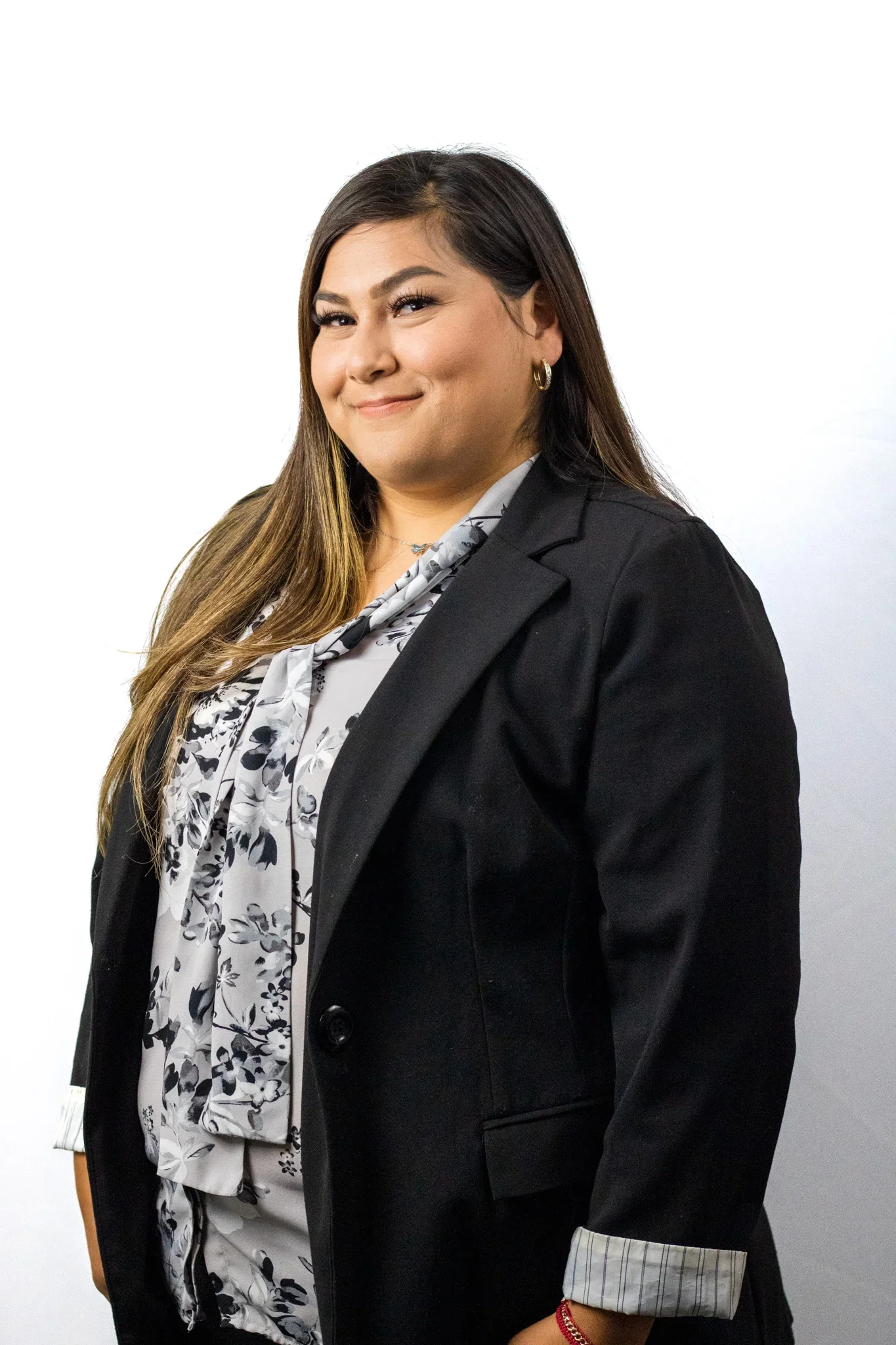 A woman with long brown hair, wearing a black blazer, white blouse with a black floral pattern, and gold hoop earrings, stands against a plain white background, smiling slightly.