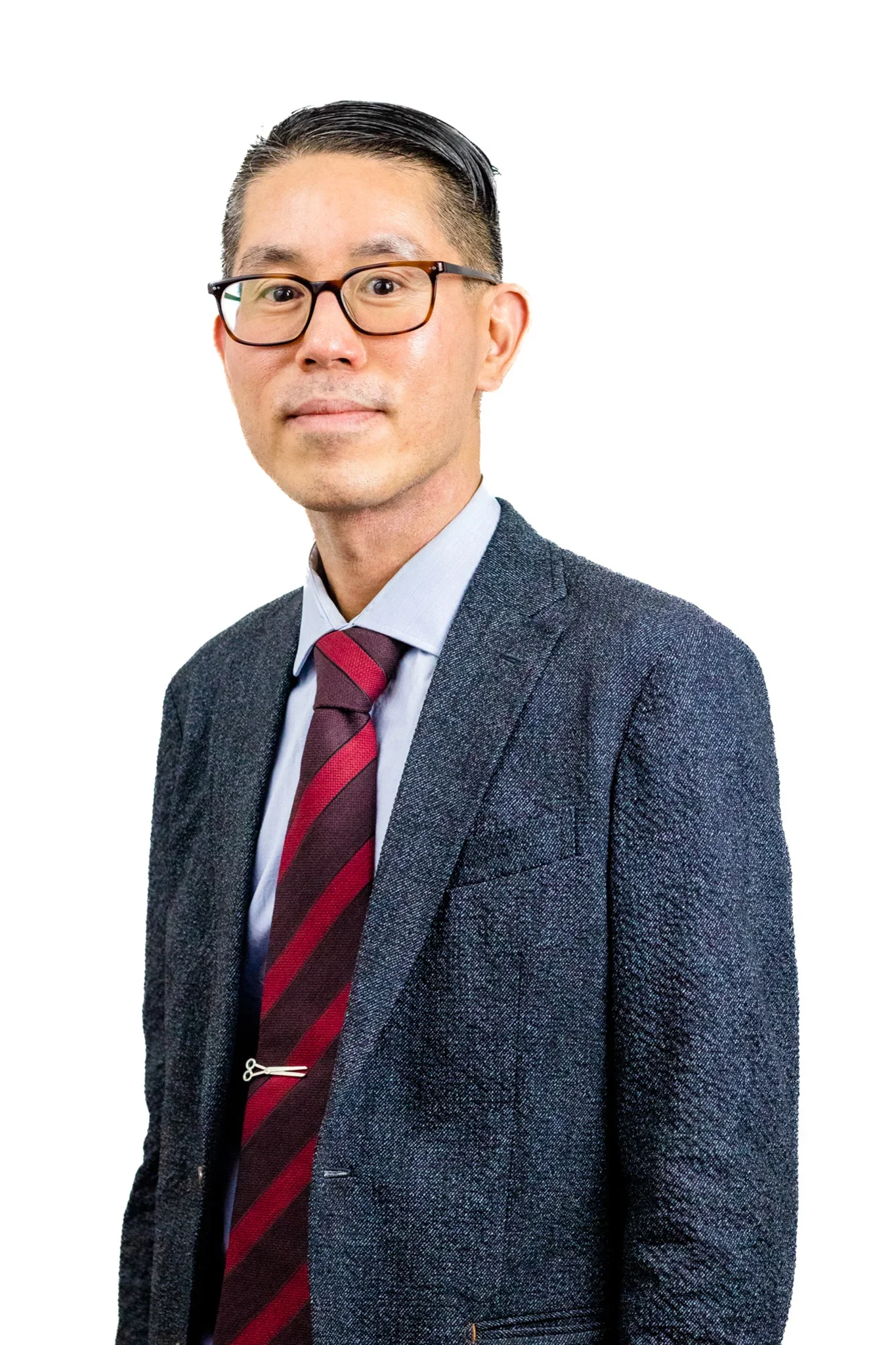 A man with short black hair, glasses, and light skin wearing a dark gray blazer, a light blue shirt, and a red and navy striped tie, posing against a plain white background.