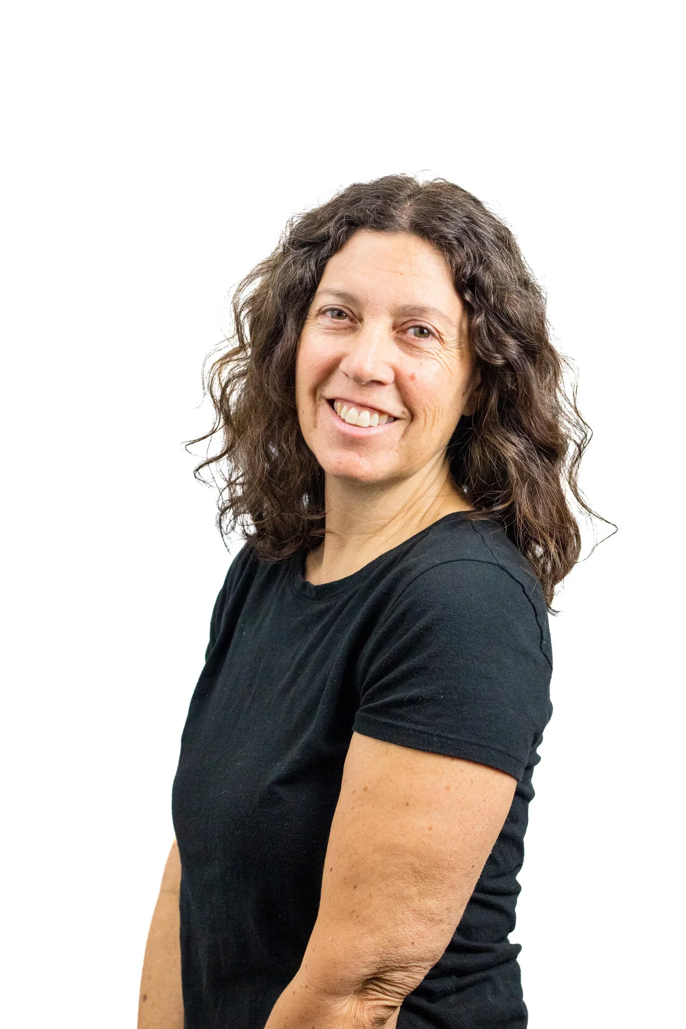 A woman with curly brown hair, wearing a black t-shirt, smiling, with her arms crossed against a white background.