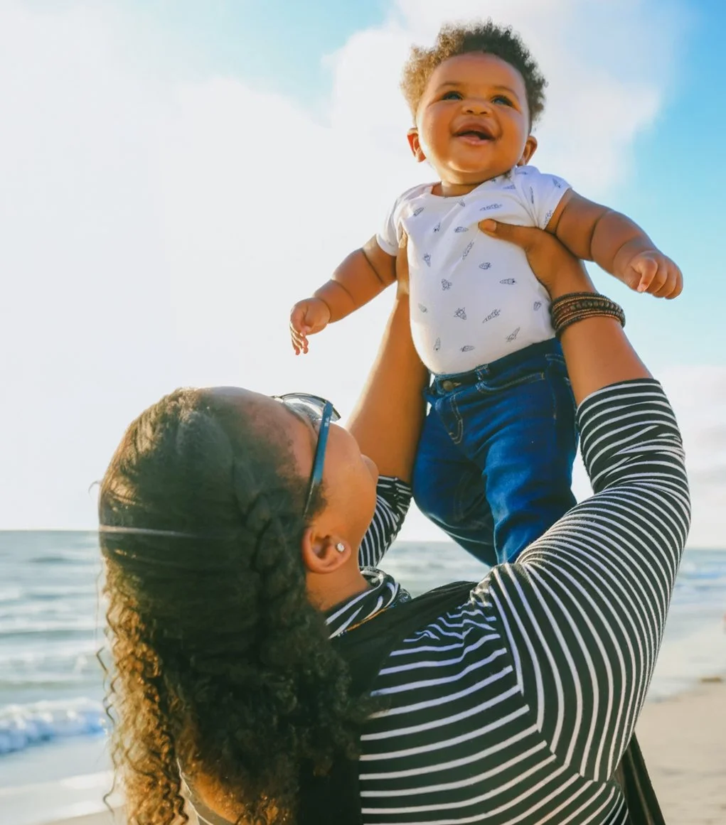 A woman with curly hair, wearing glasses and a black-and-white striped shirt, lifting a laughing baby with curly hair, wearing a white shirt and jeans, at the beach during sunset.