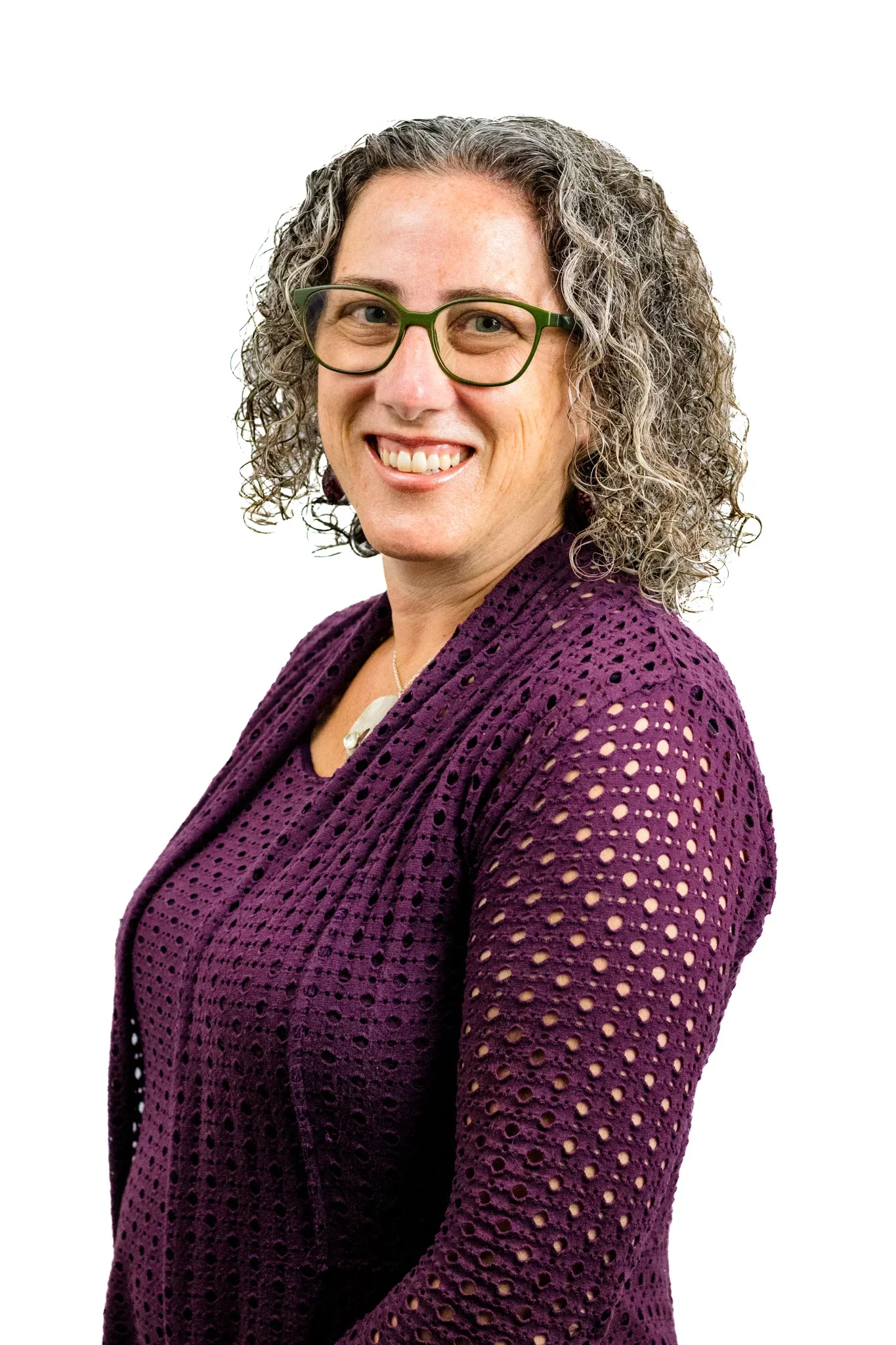 A woman with short curly gray hair, wearing green glasses and a purple crocheted cardigan, smiling at the camera against a white background.