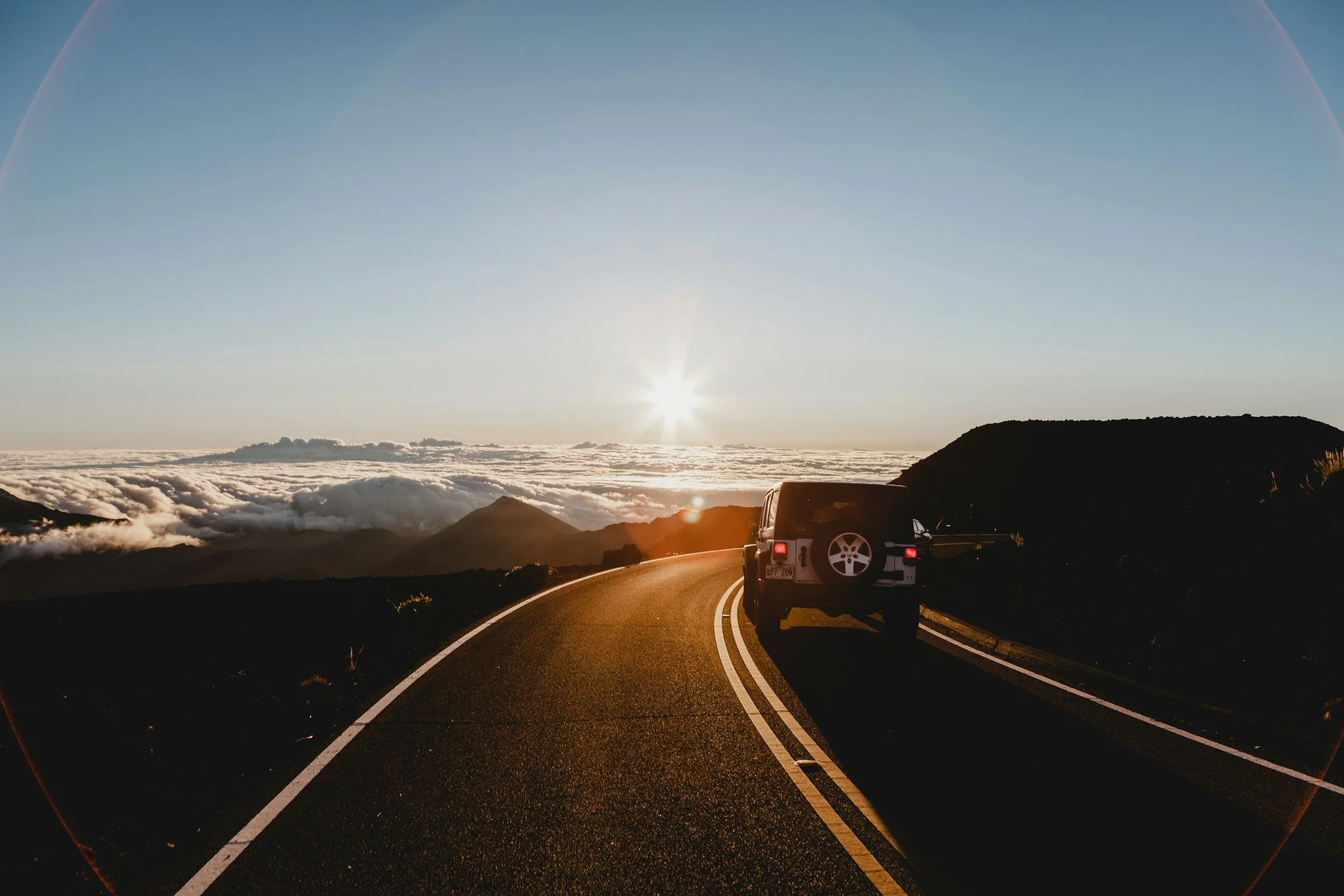 A black SUV driving on a winding mountain road at sunset, with clouds below and a visible rainbow against a clear sky.