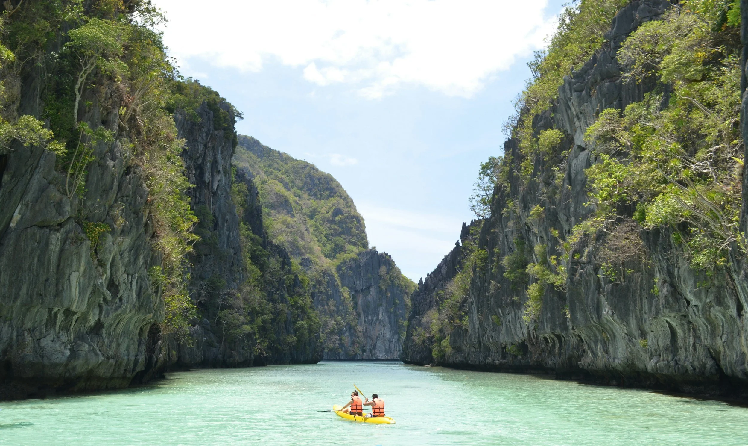 Two people kayaking on a turquoise river between high, steep, rocky cliffs with green foliage, under a partly cloudy sky.