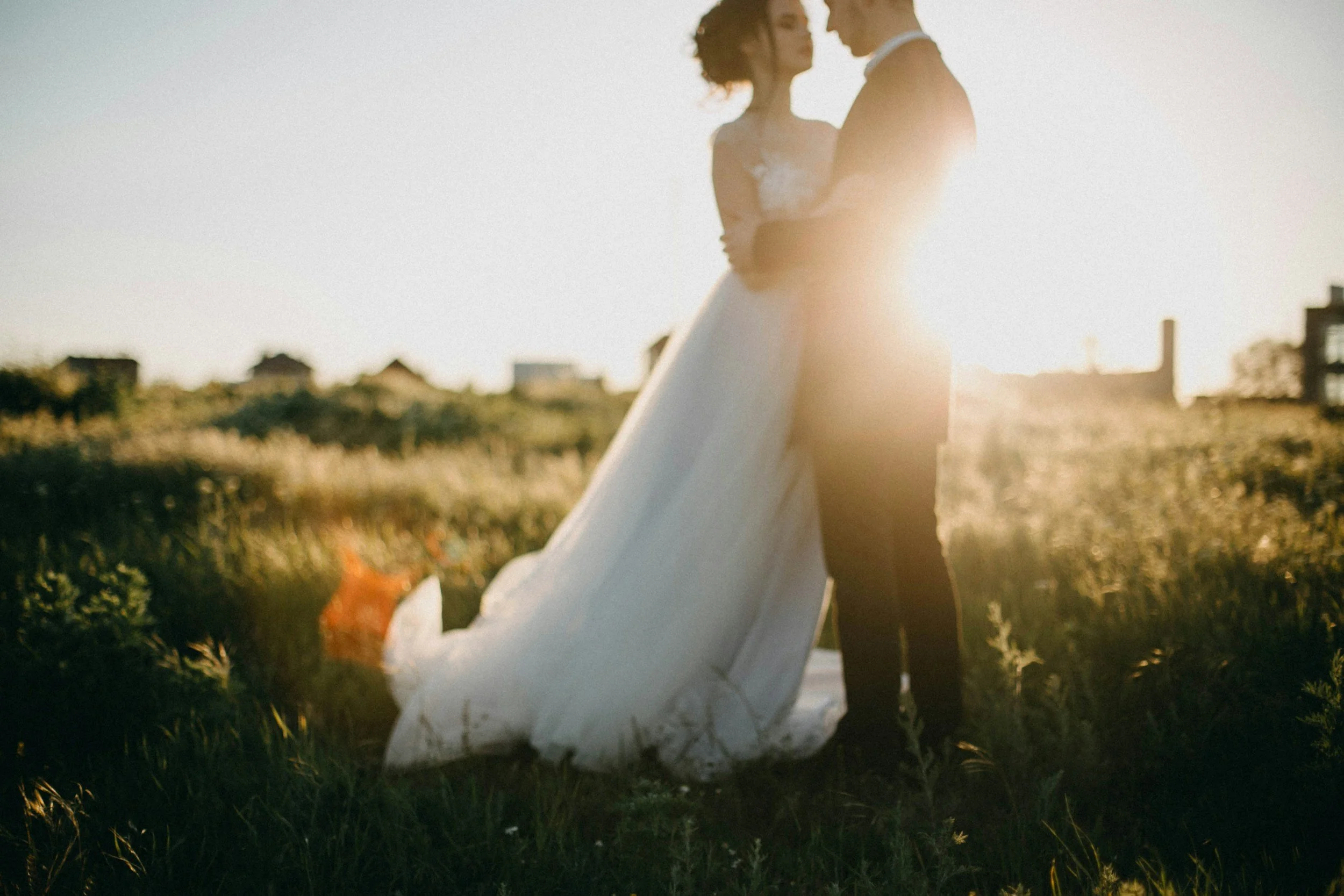A couple standing in a field during sunset, facing each other closely, with the woman in a white dress and the man in a dark suit, with warm sunlight backlighting them.