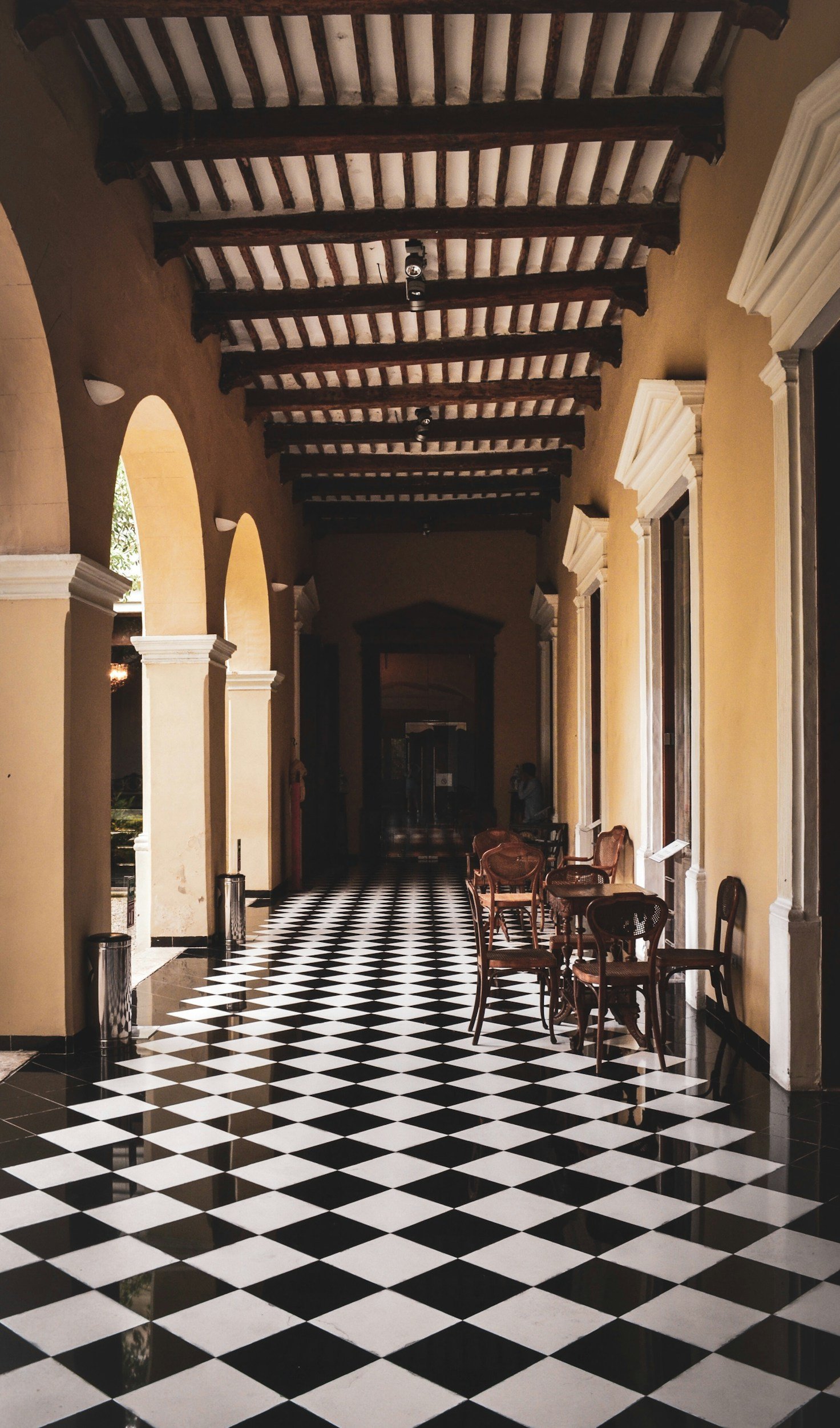 Elegant corridor with a black and white diamond patterned tile floor, yellow walls with white trim, arched doorways, and a wooden ceiling with exposed dark beams. Several chairs and small tables are positioned along the corridor, and sunlight streams in from outside.