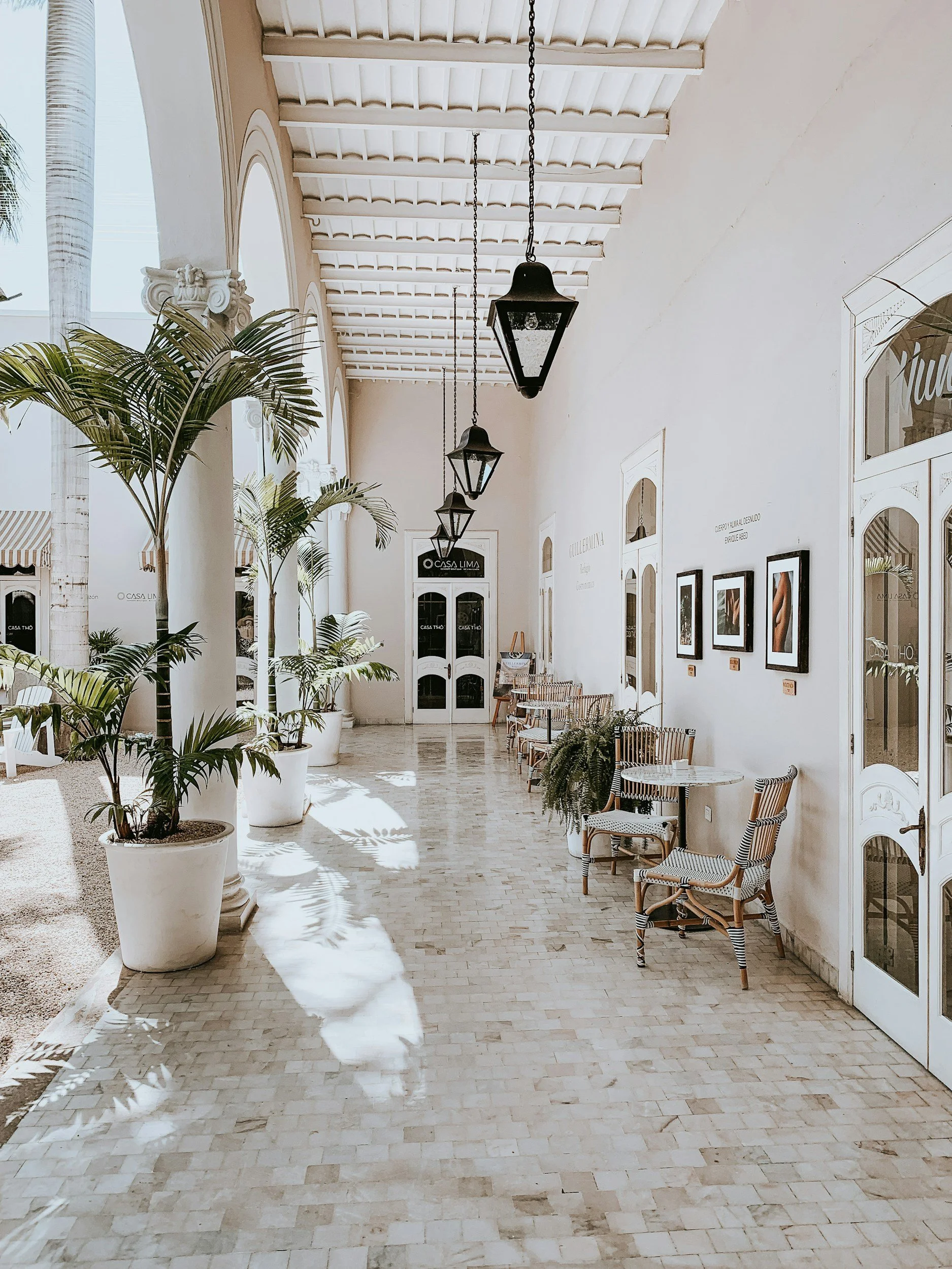 An outdoor corridor with beige tiled flooring, white walls, and hanging black lantern-style lights. There are potted plants, including palms, and seating with chairs against the wall. The space is bright, with natural light coming from the left. There are framed pictures on the wall and arch-shaped windows and doors.