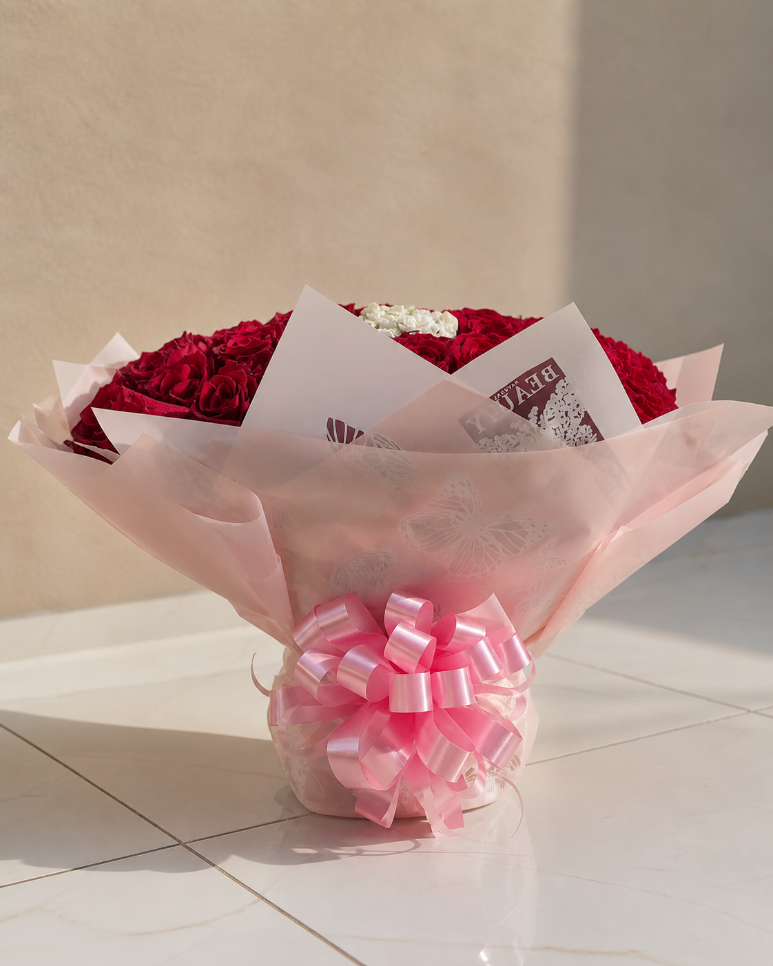Ramo de flores con rosas rojas, blancas y rosas, decorado con papel trasparente y lazo rosa, colocado sobre una superficie de baldosas.