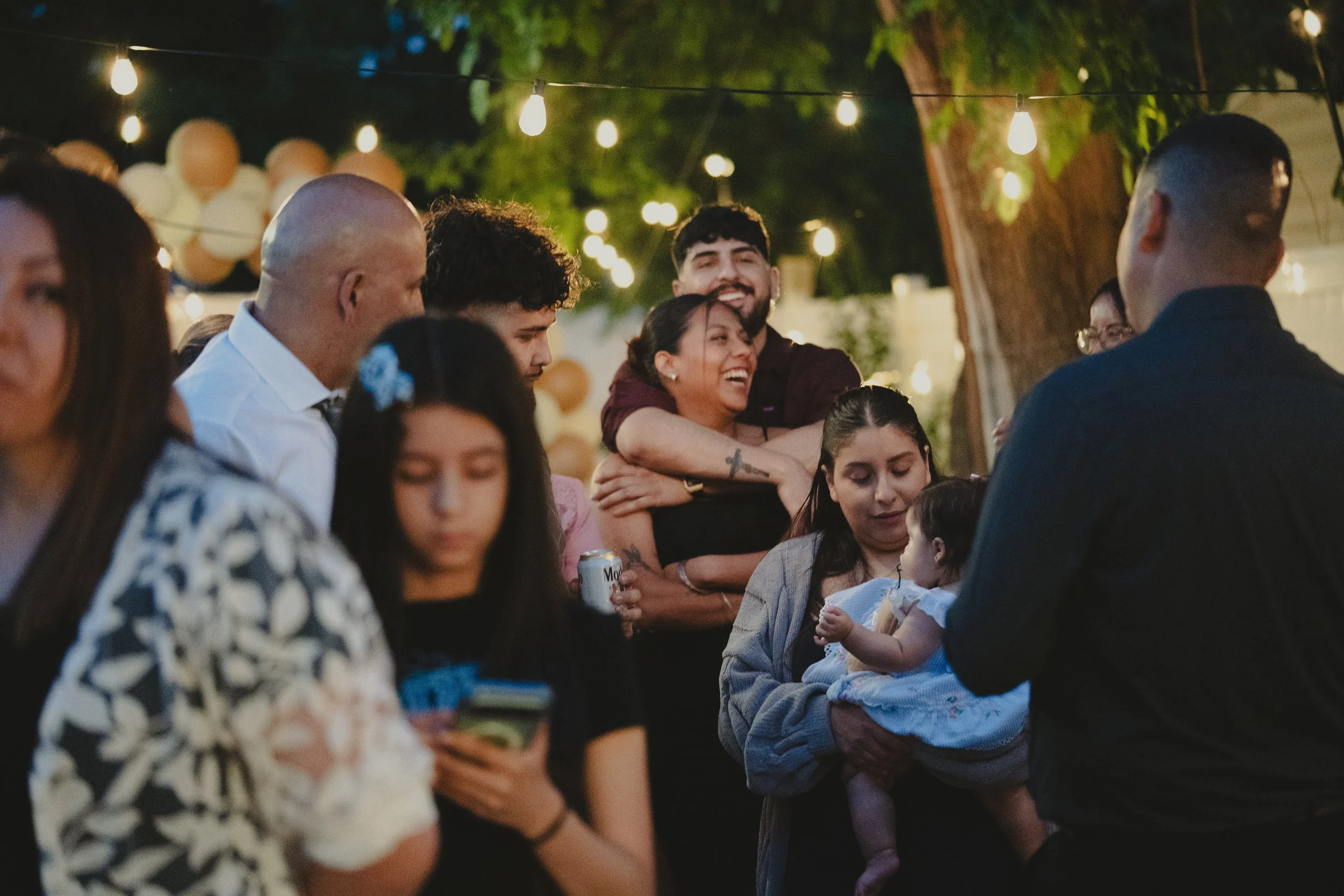 People gathered outdoors at night under string lights, socializing and smiling, including adults and children, with balloons and trees in the background.