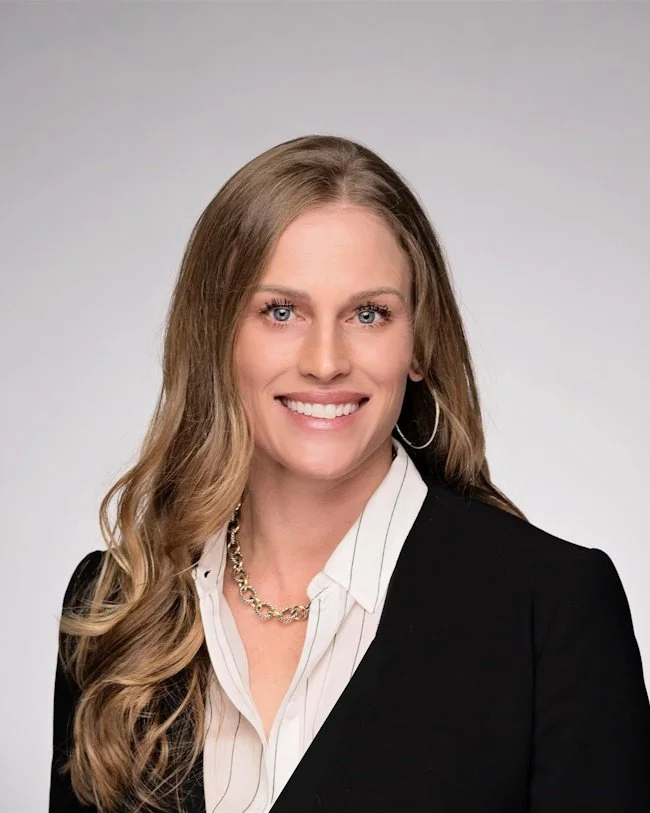 Headshot of Erin Mcerlane, relocation specialist at Latreuo Real Estate. Professional woman with long wavy blonde hair, blue eyes, smiling, wearing a black blazer, white shirt, gold chain necklace, and hoop earrings, against a light gray background.