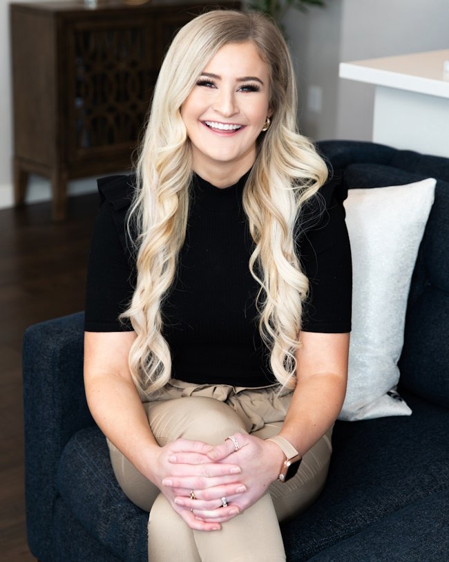 Headshot of Kaitlin Tookey Palmer, realtor with Latreuo Real Estate. A woman with long blonde hair sitting on a dark blue couch, smiling, wearing a black top and beige pants, in a well-lit room.