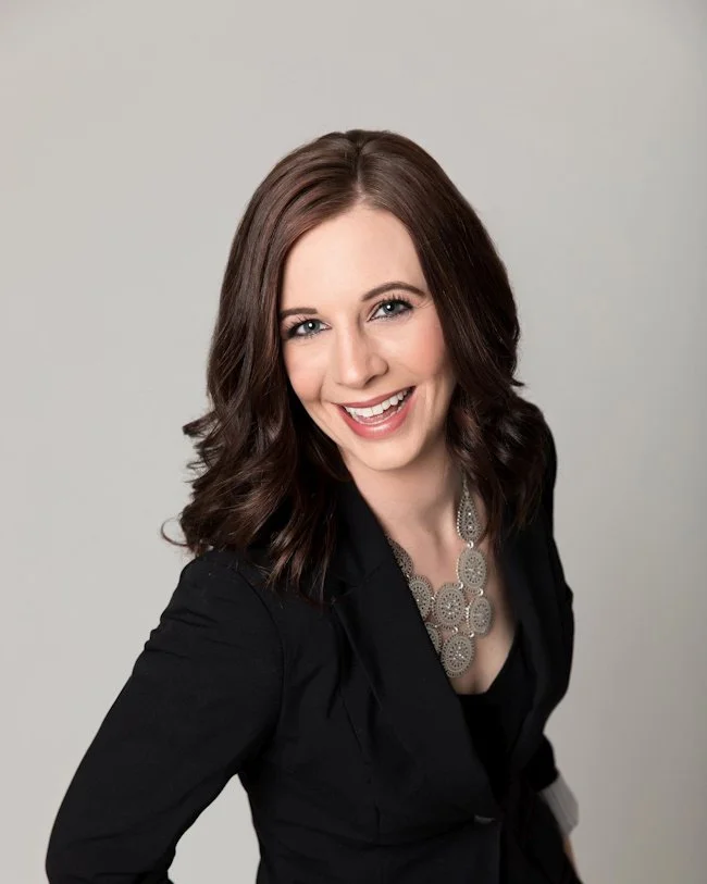 A woman with shoulder-length brown wavy hair, smiling, wearing a black blazer and a large silver necklace, against a plain light gray background.