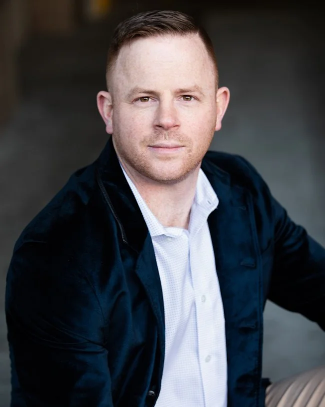 Headshot of Casey Wilson, owner, founder, and realtor with Latreuo Real Estate. Short light brown hair, blue eyes, and light skin, wearing a white collared shirt and dark jacket, sitting outdoors with a blurred background.