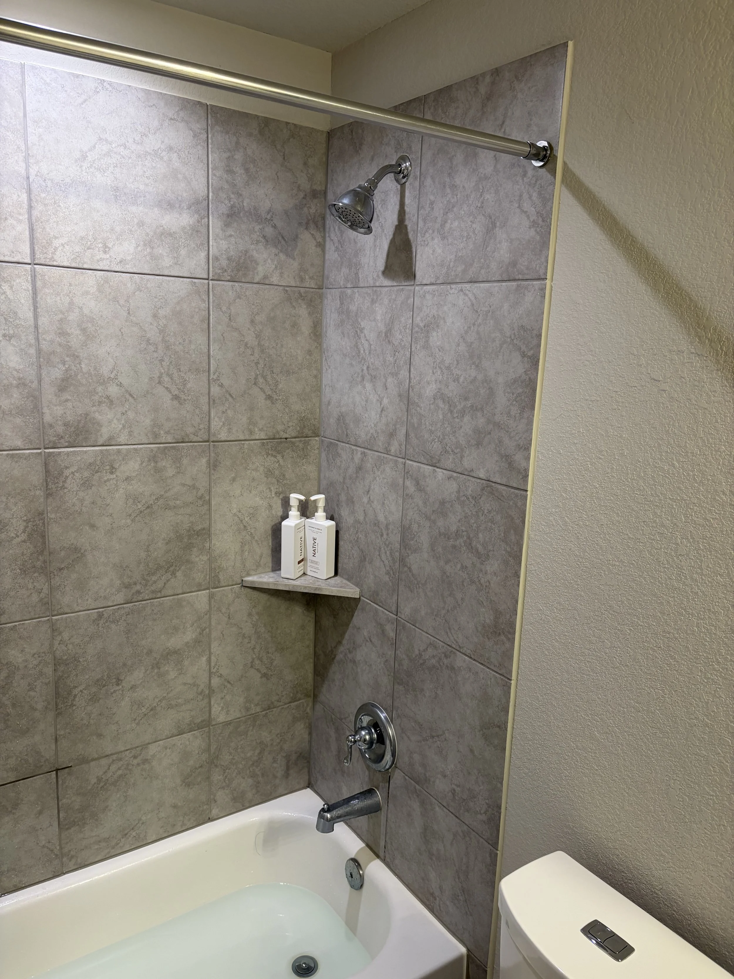 Bathroom shower and bathtub with beige tiled wall, silver showerhead, shower faucet, two bottles of shampoo, and part of a toilet.