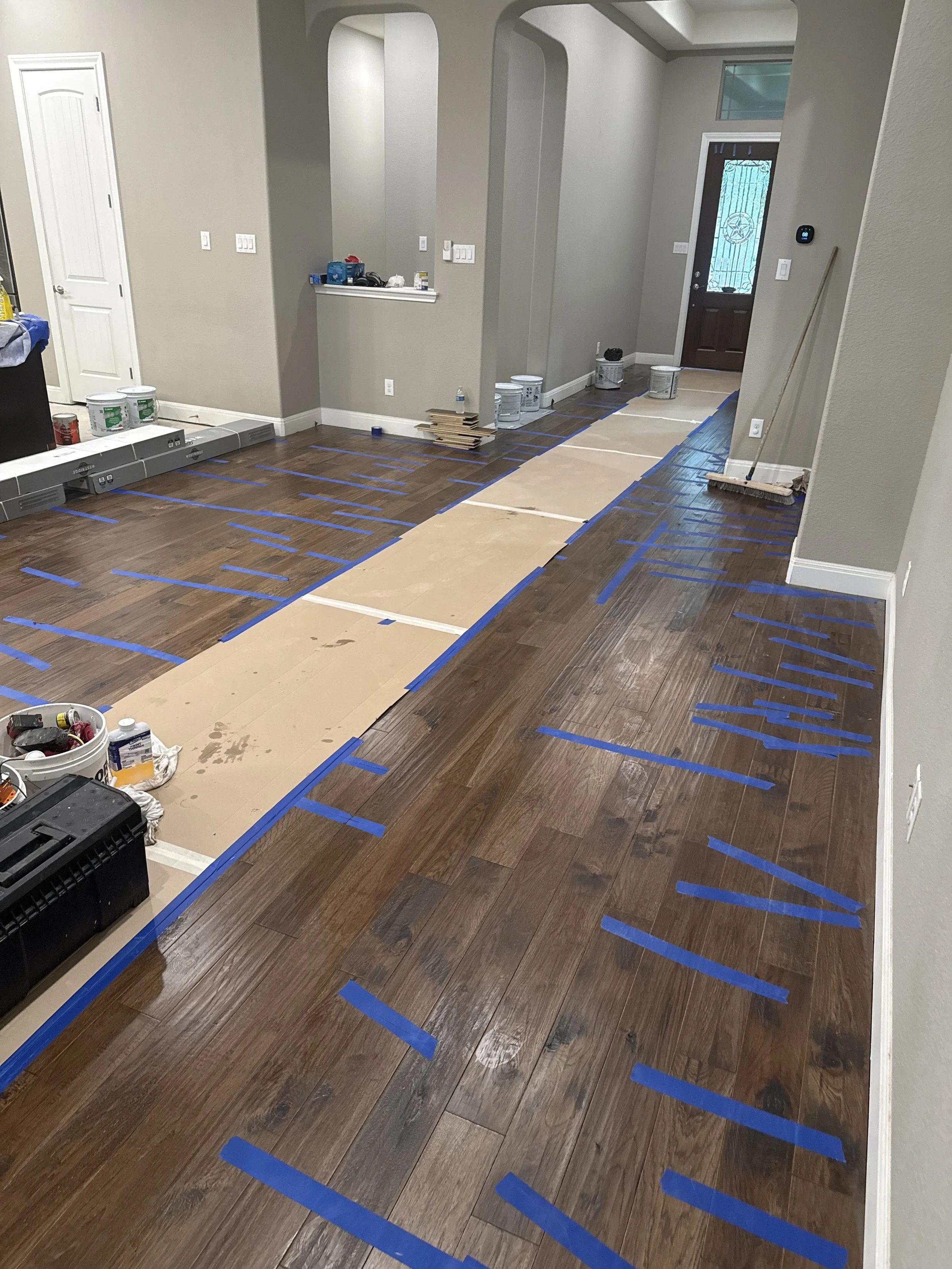 Living room with partially installed hardwood flooring, blue painter's tape marking sections, tools, and paint cans on the floor, and a front door with decorative glass window.