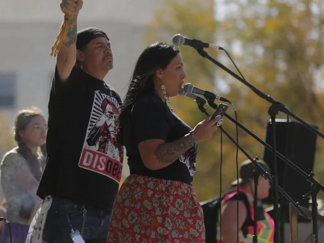 Woman with dark hair reading from a phone at a microphone at an outdoor protest, man behind her raising a fist, other protesters in the background.