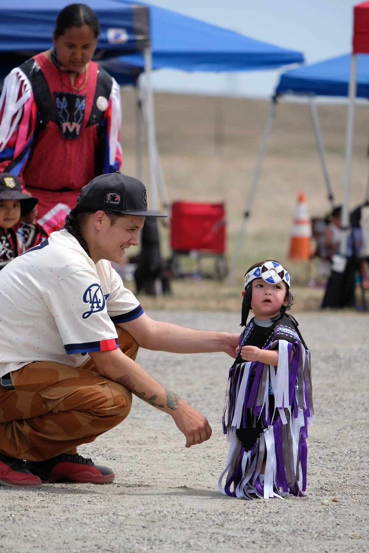 A man kneeling and smiling at a young girl wearing a costume with purple, white, and black streamers, with a headband and day of the dead face paint, at an outdoor event with tents and other people in the background.