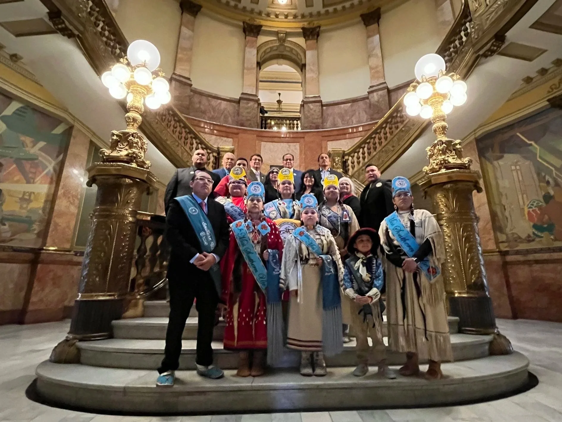 A group of children in Scout uniforms with adults, standing together on a staircase inside a grand, ornate building with marble pillars, gold accents, and large lamps, posing for a photo.