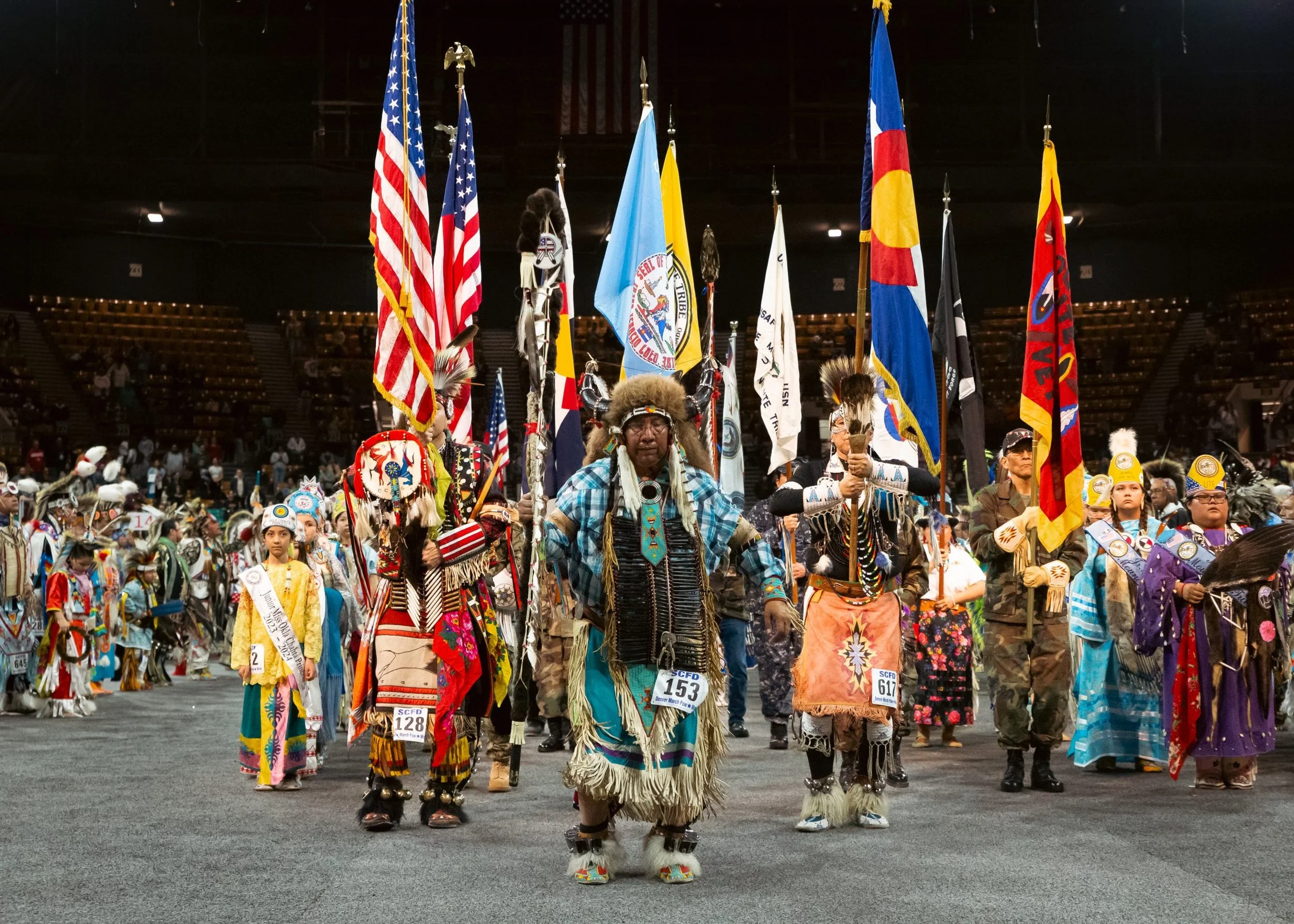 A group of Native Americans dressed in traditional clothing holding flags at a cultural event or parade inside a large indoor arena.