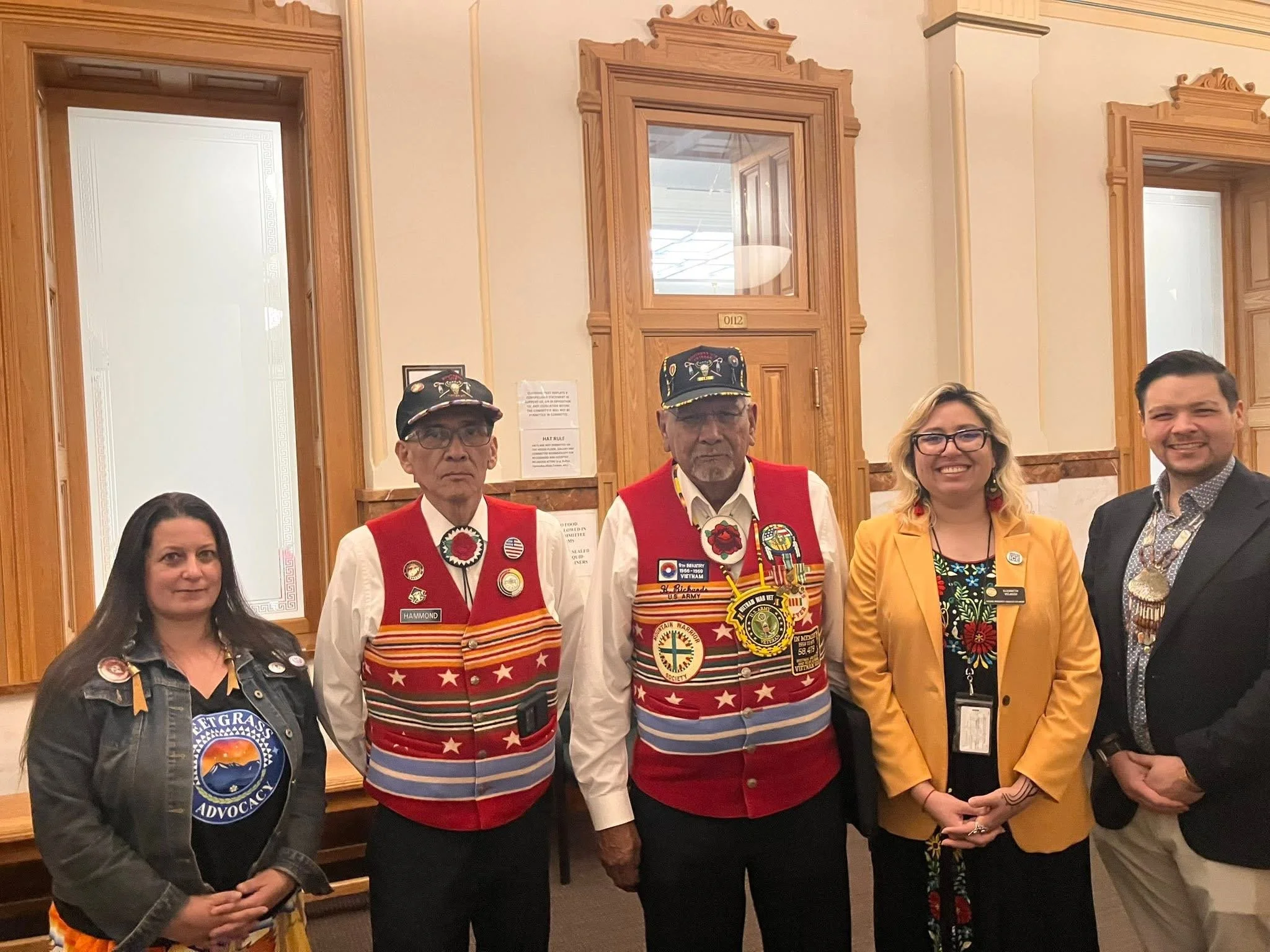 Group of five people standing indoors in front of wooden-framed windows, wearing organization or event badges and clothing, with some dressed in vibrant, decorated outfits featuring patches and pins, smiling and posing for the photo.