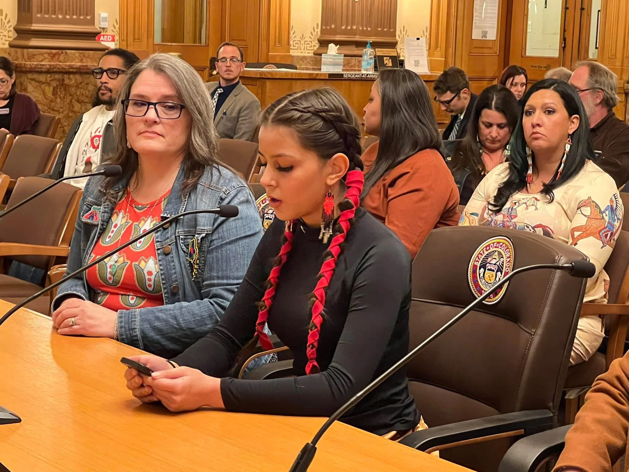 A group of diverse people sitting at a long wooden table in a formal meeting or hearing room with wood paneling and decorative features. Many are looking down at their phones, others are looking forward or to the side. A woman in the foreground has long black hair with red braids and earrings, wearing a black shirt. The woman next to her has gray hair, glasses, and is wearing a denim jacket over a patterned shirt. In the background, others are seated with some wearing glasses, suits, or traditional attire.