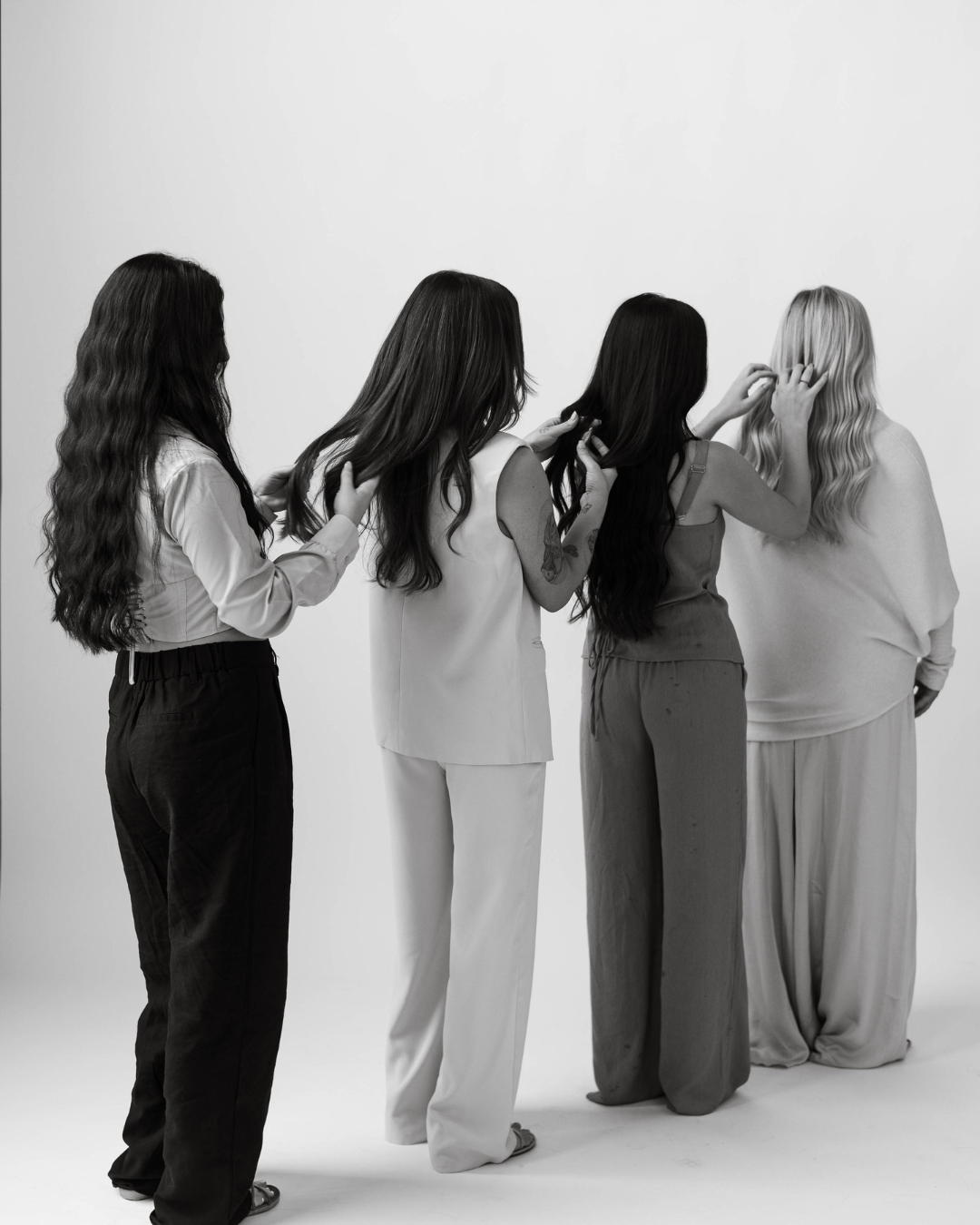 Four women with long hair, standing in a line, styling each other's hair, facing away from the camera against a plain wall.